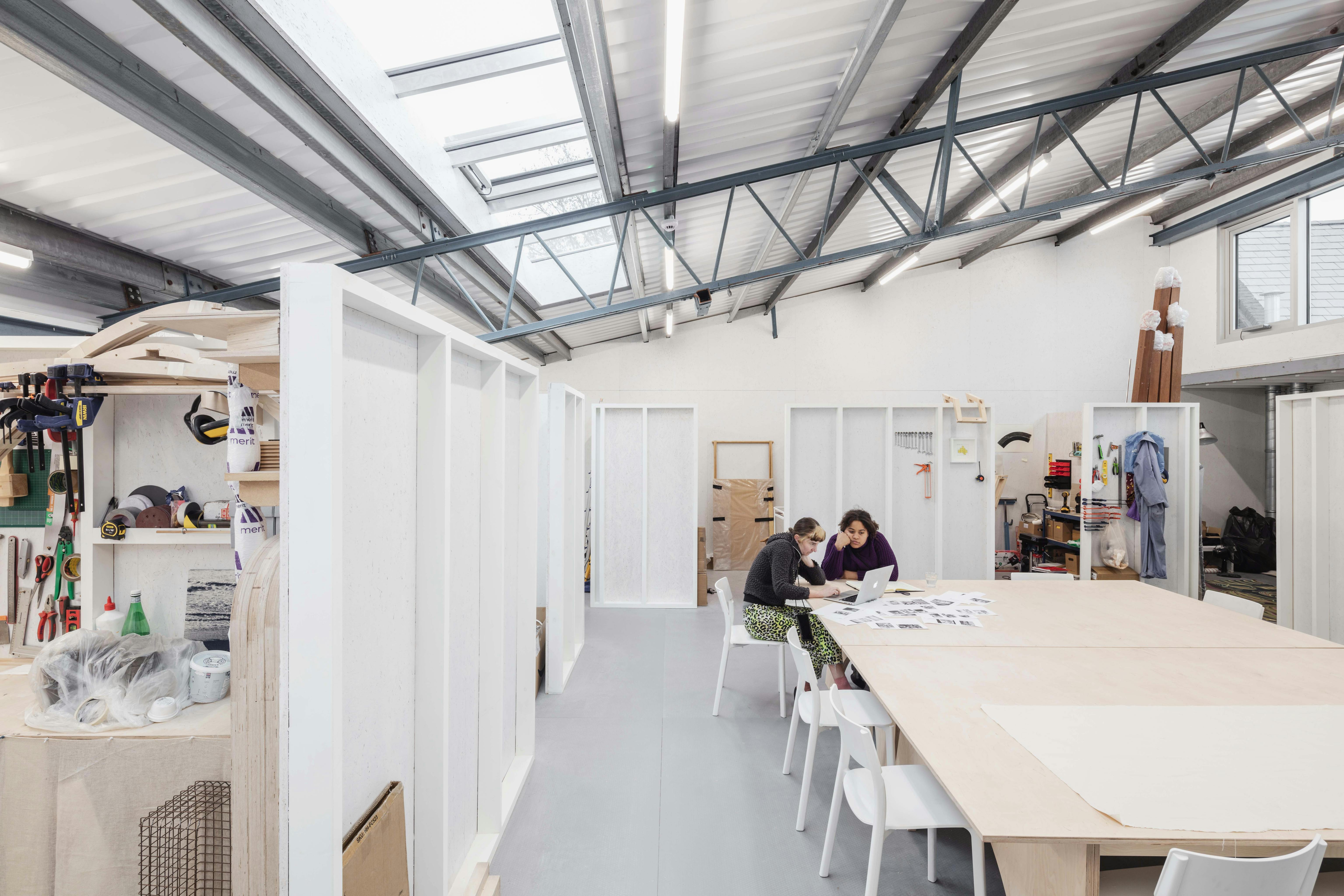 A bright, spacious studio with white walls, large windows and exposed beams. Two people sit at a large table covered with papers, while art supplies and materials are neatly organised on shelves in the background.