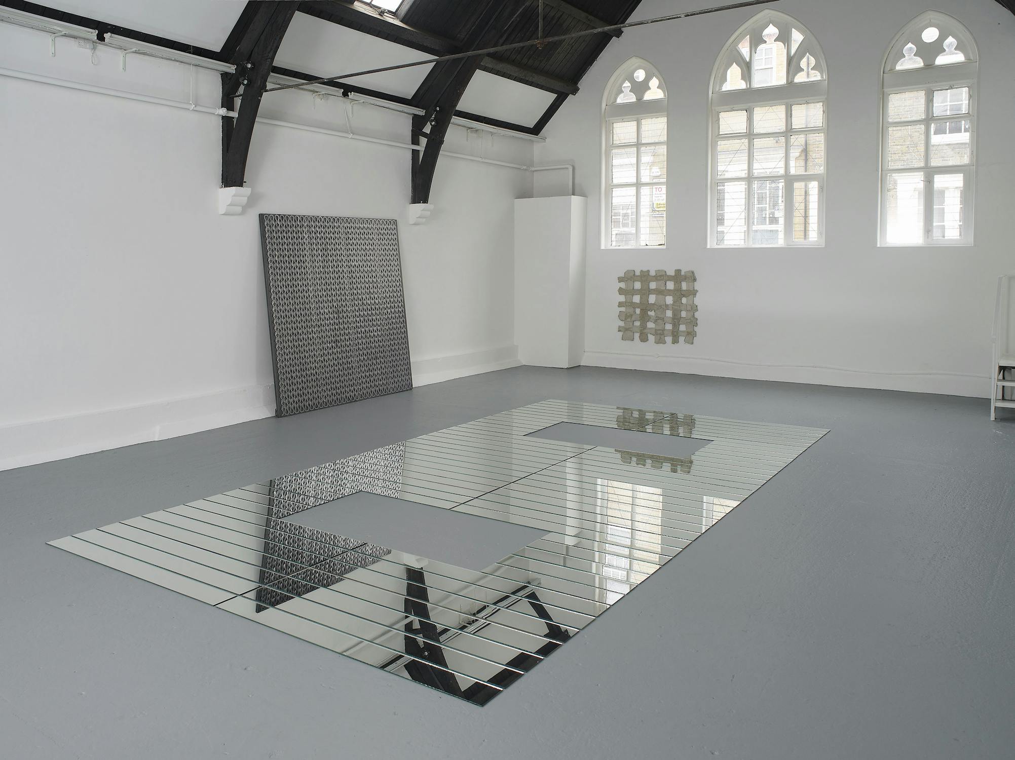 A bright, high-ceilinged gallery features a large mirrored tile installation on the floor with two square voids. Against the white walls are a metal grid panel and a grid of beige blocks. Arched windows let in natural light, illuminating the minimalist scene.