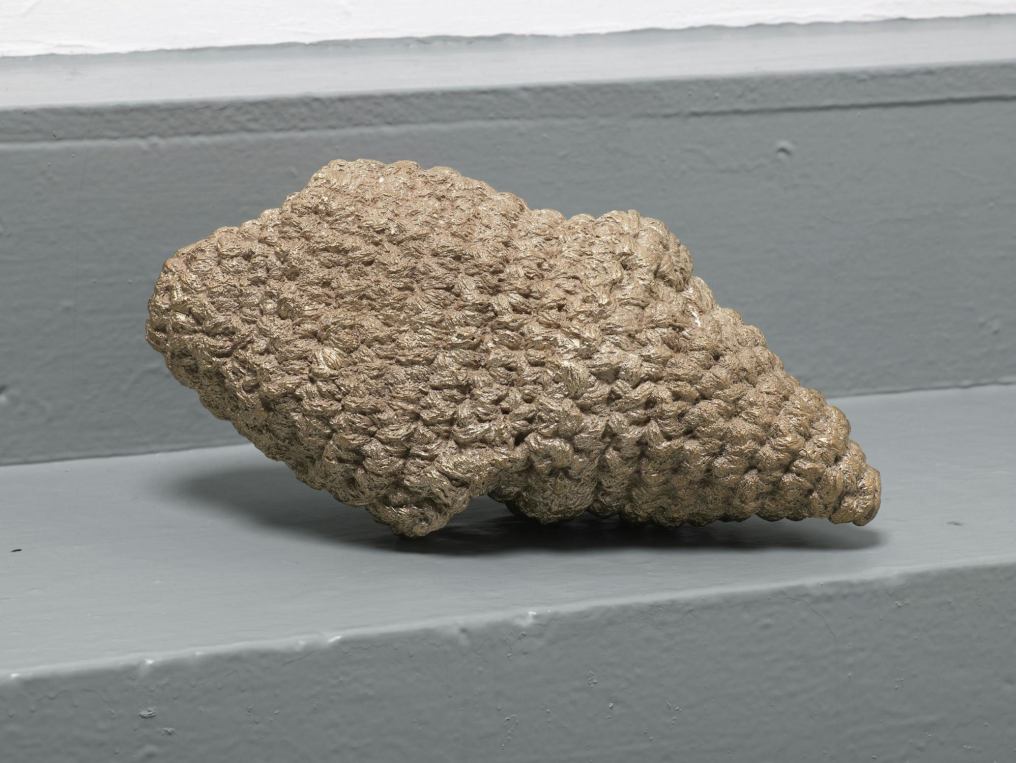 A textured, cone-shaped object with a rough, bumpy surface rests on a smooth, grey-painted step. The object is beige-brown, resembling a pine cone or wasp nest, and is positioned slightly diagonally in the image.