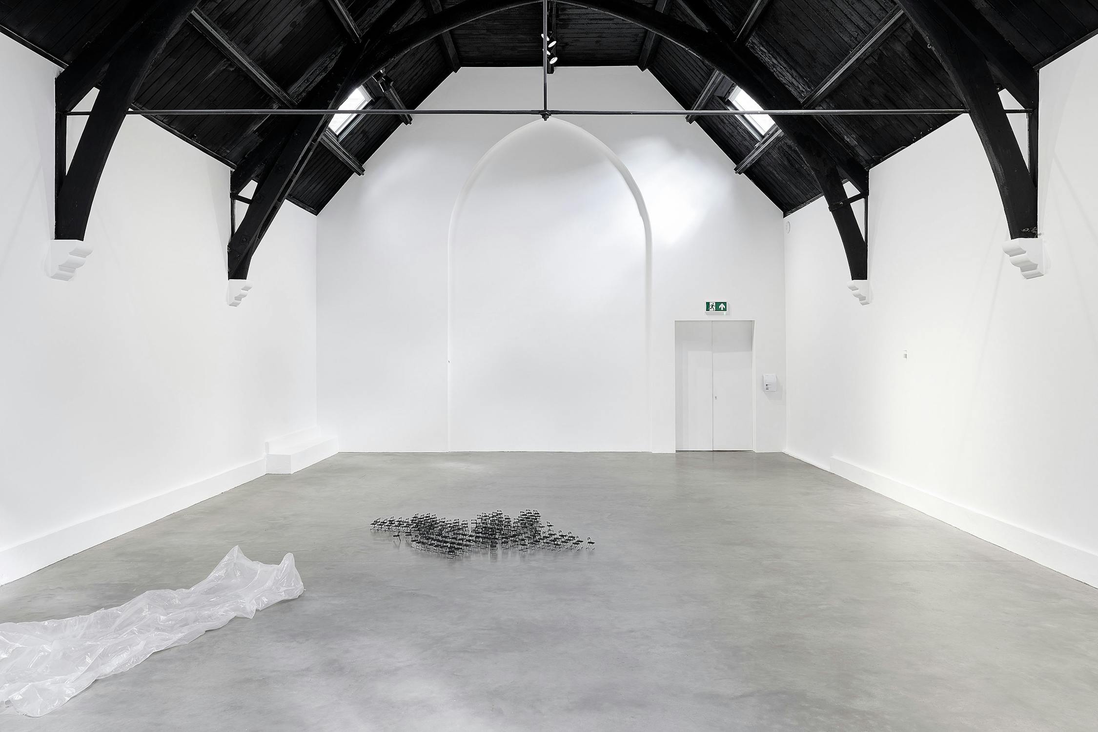 A minimalist art gallery with high, dark wooden ceilings, white walls, and a grey concrete floor. A translucent sheet lies on the left floor, and a small cluster of black objects is centred in the room.