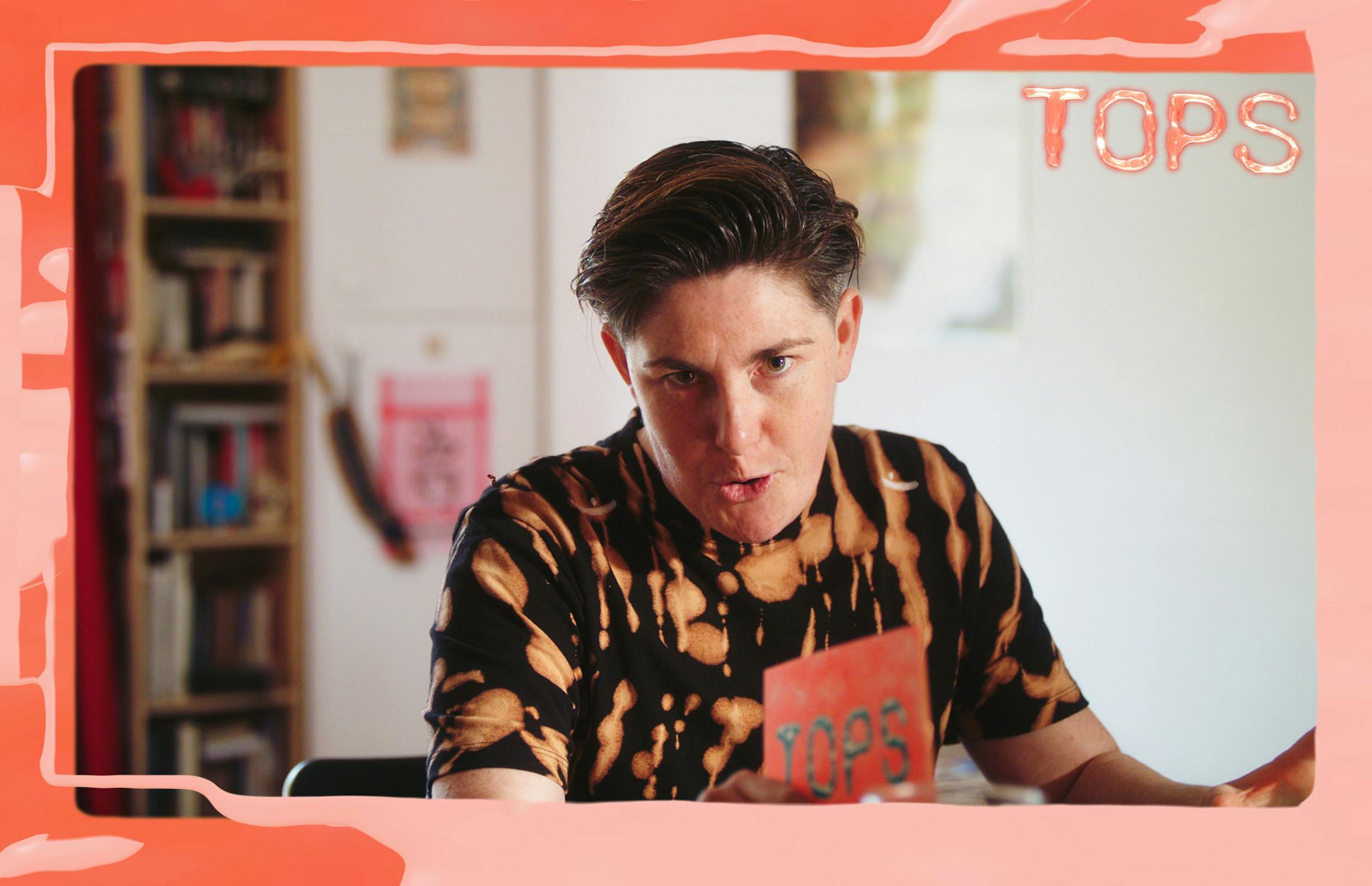 A person with short, dark hair wearing a black and orange tie-dye shirt holds a red card labeled "TOP5." They sit indoors, framed by a graphic border with "TOPS" text in the corner. Shelves and posters are visible in the background.