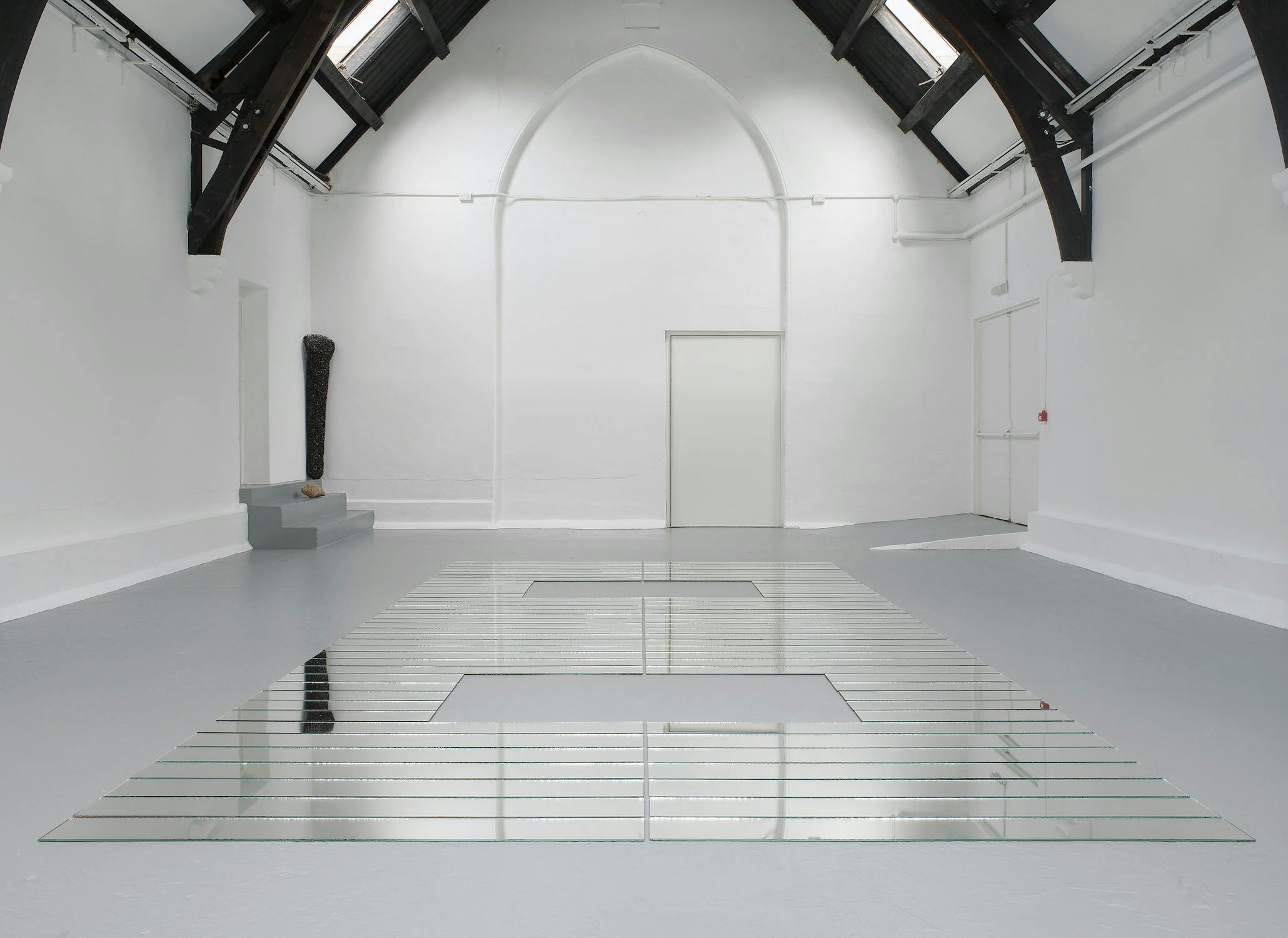 A minimalist, modern gallery space with white walls and a high arched ceiling features a grid of reflective glass panels arranged on the floor and a black, textured sculpture on a small grey platform in the back left corner.