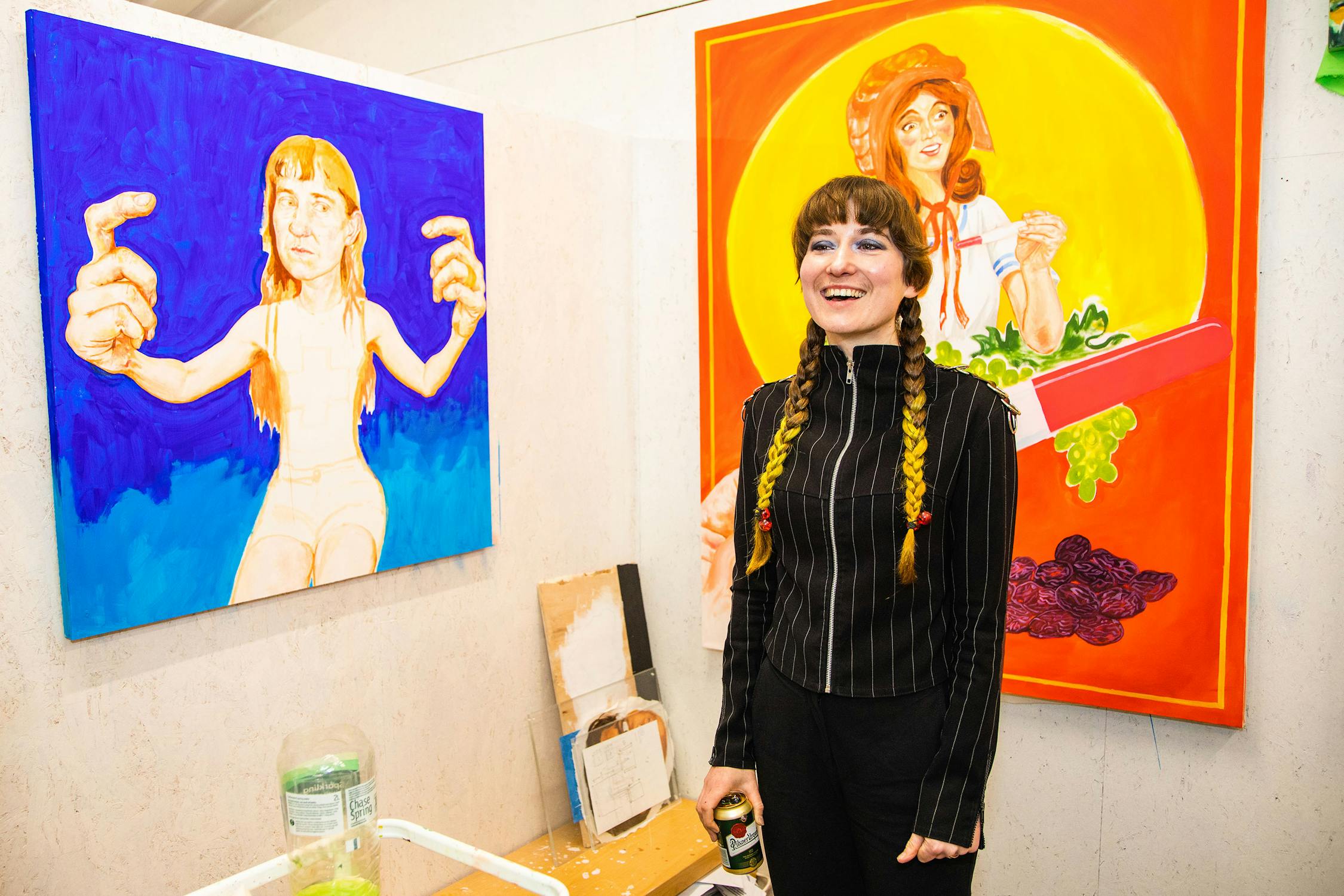 A smiling person with long braided hair stands in front of colorful paintings, one blue and one orange, in an art studio. The paintings feature abstract human figures with exaggerated hands and playful expressions.