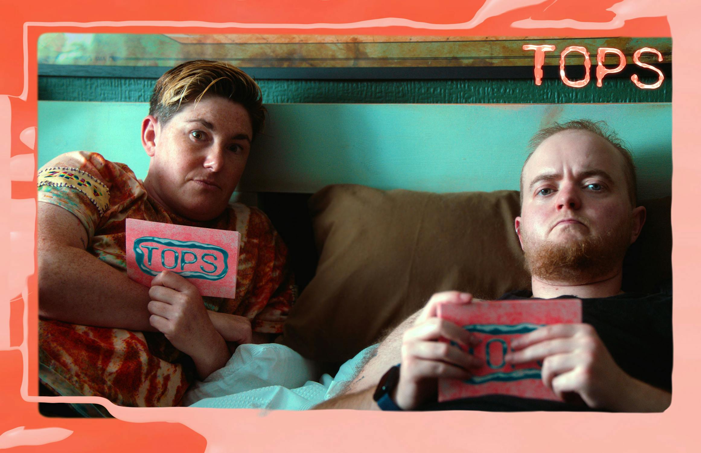 Two people sit on a bed holding cards that say "TOPS." Both have serious expressions. The image has a coral-coloured border with the word "TOPS" in the top right corner.