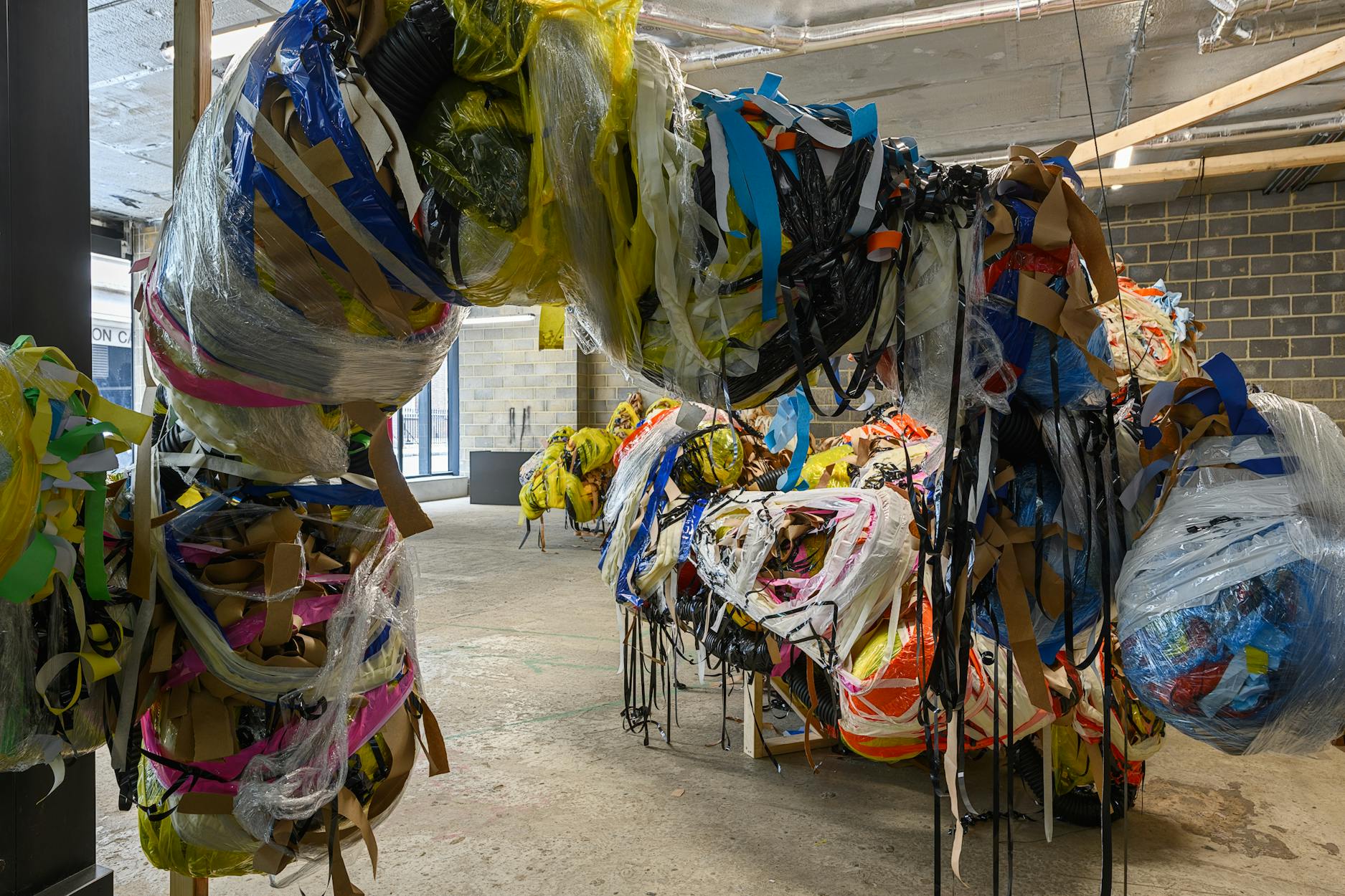 A large abstract sculpture by Nnena Kalu made of bundled plastic, tape, fabric, and cords in various colours, dominates an industrial gallery space with bare concrete floors, exposed pipes, and brick walls. The materials are wrapped chaotically, forming organic, twisting shapes.