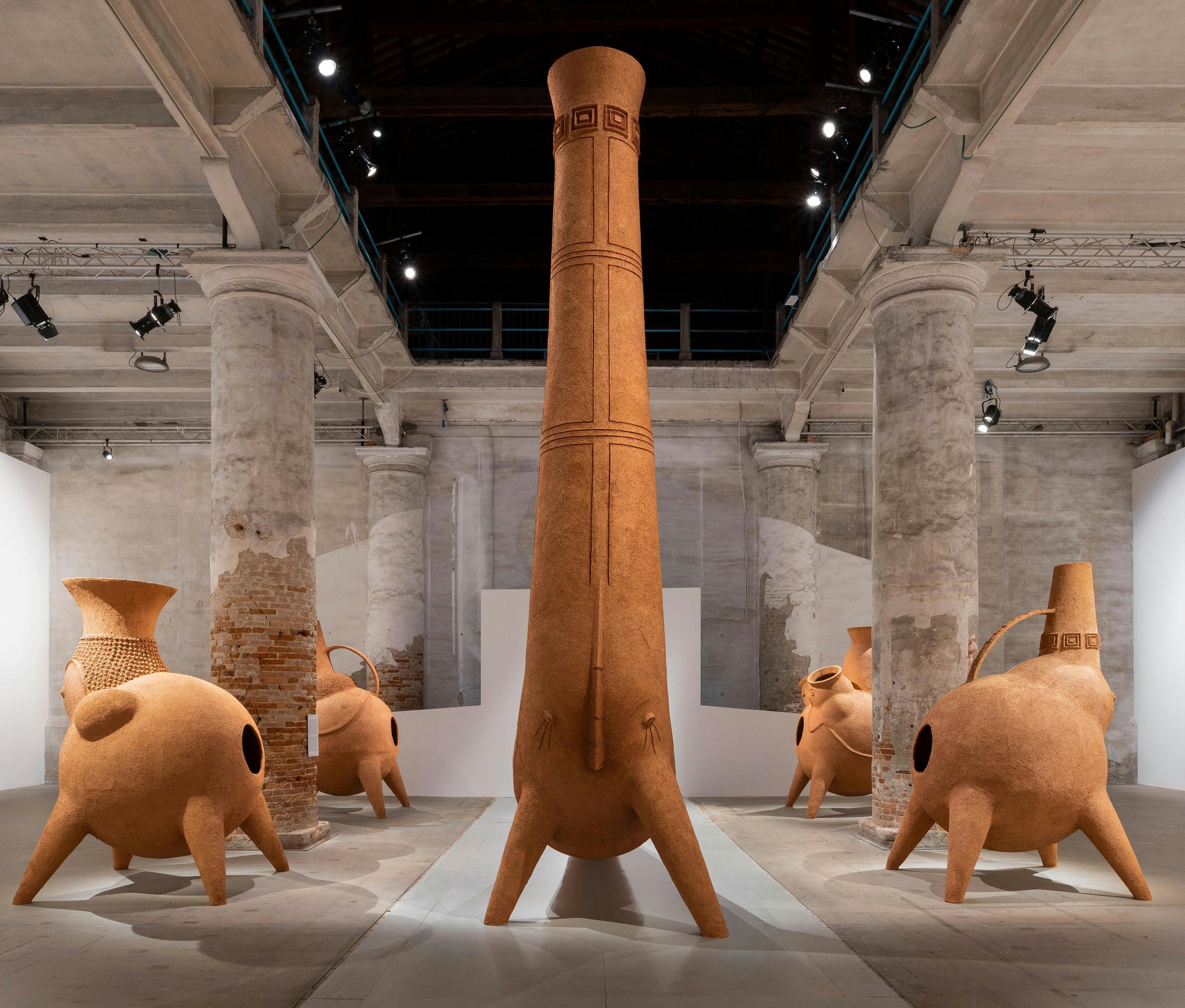 A towering vessel with legs, flanked by smaller and squatter clay ovens, in a gallery setting