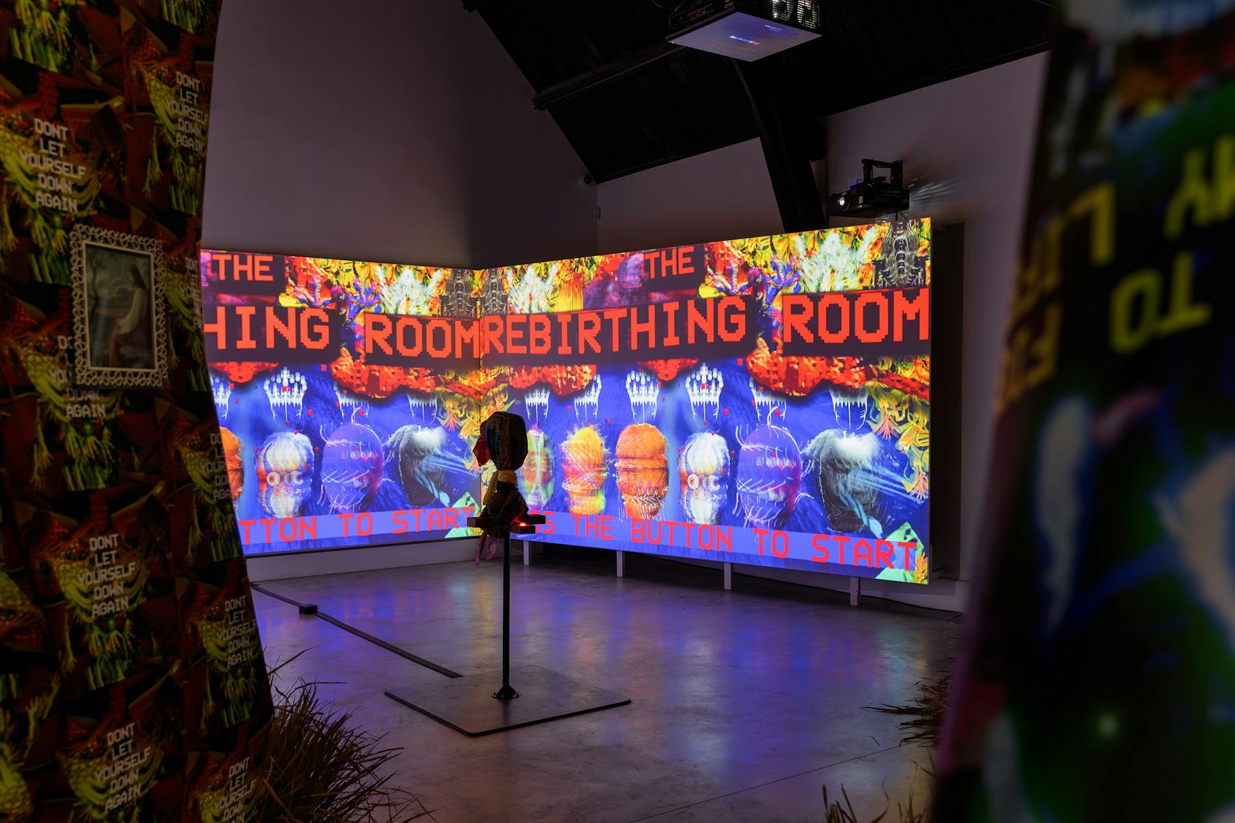 a photograph of two large wall sized screen with projections that read 'THE REBIRTHING ROOM'. In the foreground there is a hooded object on a stand.  