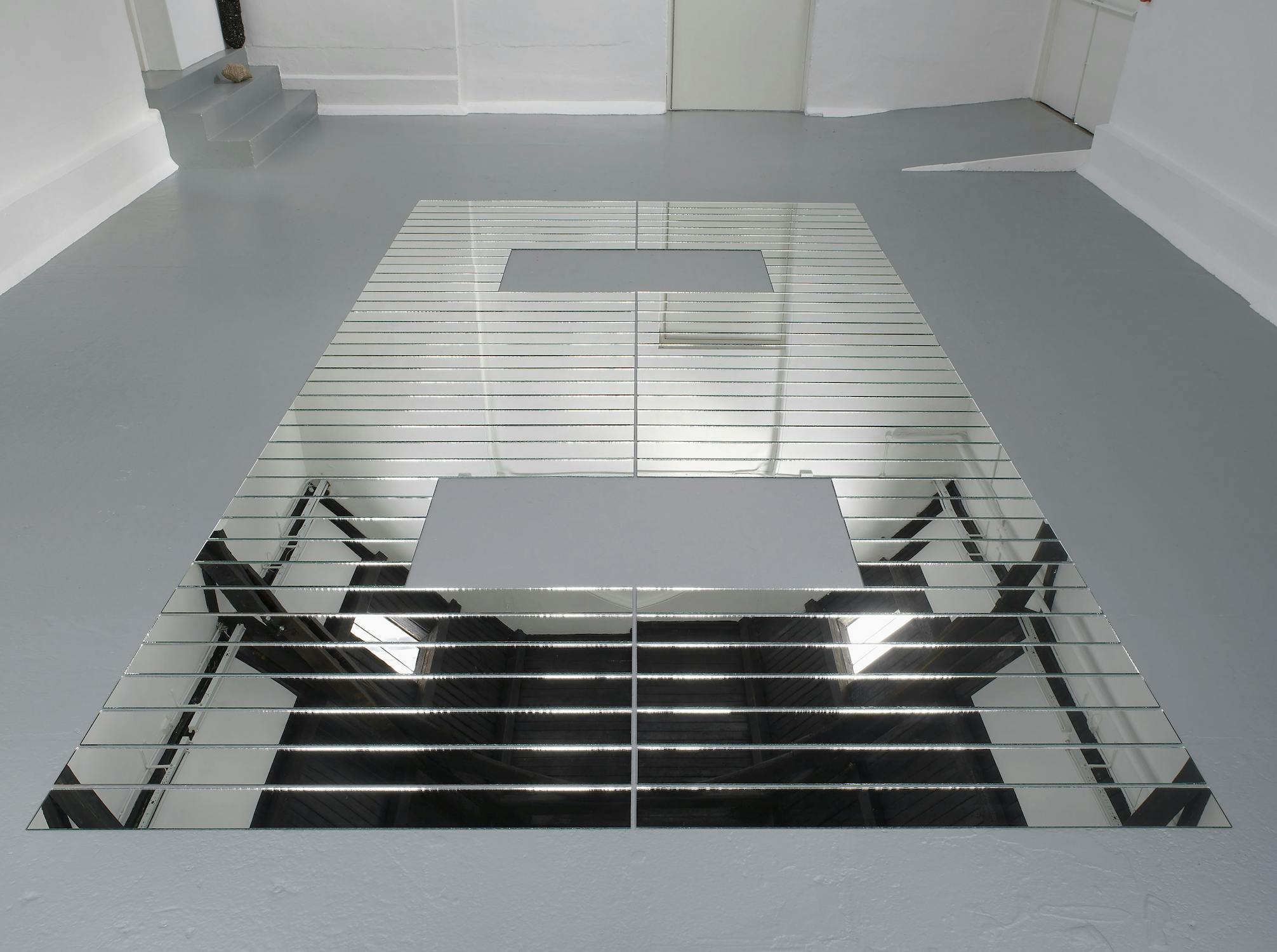 A geometric floor installation made of rectangular mirrored tiles is arranged in a symmetrical, angular pattern. Two rectangular cut-outs are in the centre, reflecting the white walls, grey floor, and room’s lighting, creating an illusion of depth.
