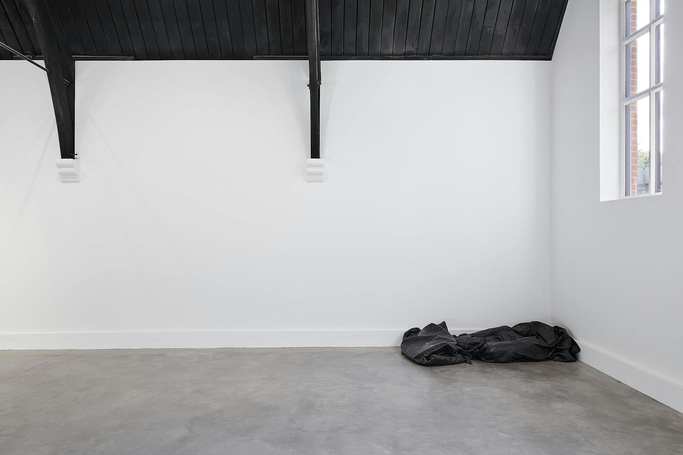 A minimal gallery space with white walls, a gray concrete floor, a black wooden ceiling, and a large window. A crumpled black leather body bag is placed in the corner near the window.