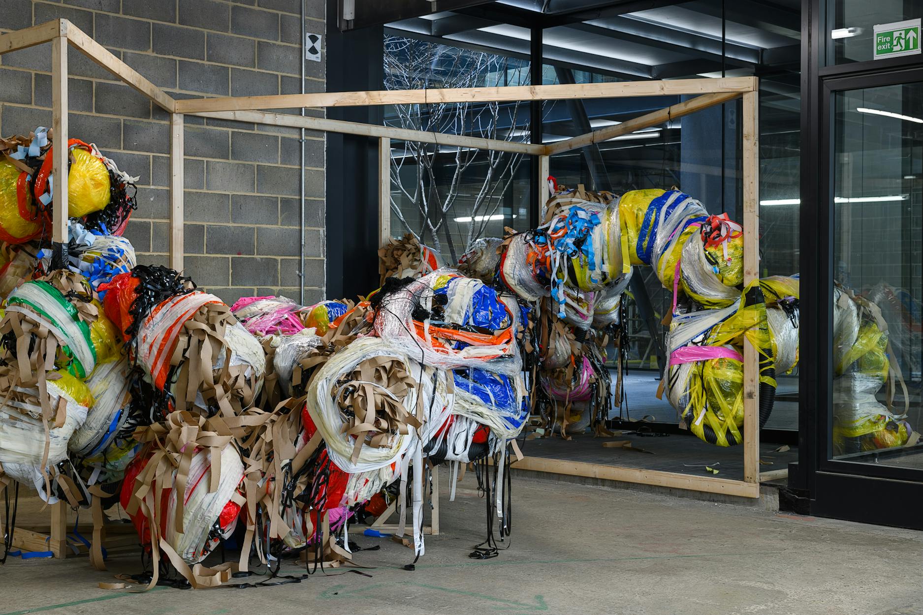 A large, abstract sculpture made of bundled, colourful plastic and paper strips sits inside a simple wooden frame structure within an industrial-style room with brick walls, glass doors, and exposed vents. The artwork spills out of the frame, appearing dynamic and tangled.