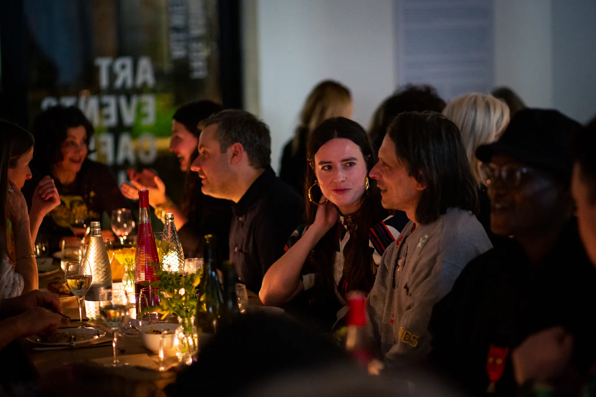 A group of people sit closely together at a warmly lit dinner table, engaged in conversation. Bottles, glasses, and candles decorate the table.