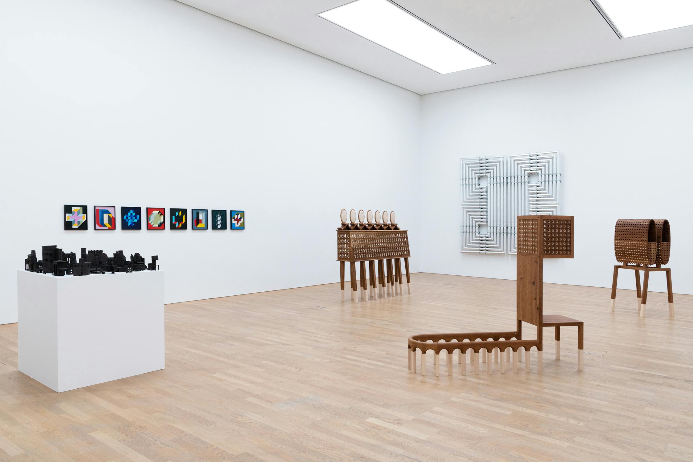 An art gallery with modern sculptures and paintings. Wooden geometric sculptures are displayed on the floor, and colorful abstract paintings hang on the white walls. Skylights illuminate the room with natural light.