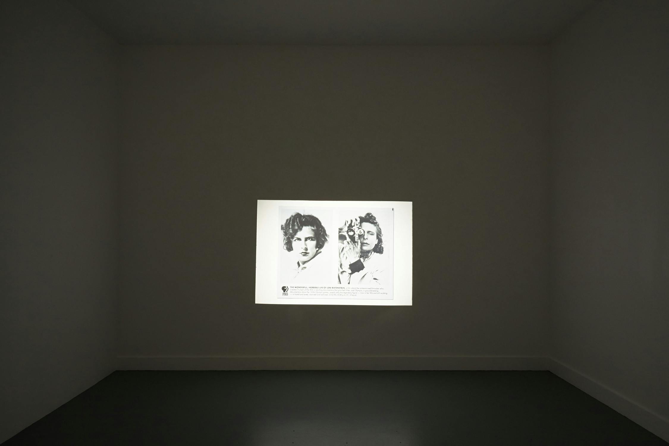 A black-and-white photo is projected onto a plain, light-coloured wall in a dimly lit room. The photo depicts two people engaged in conversation, one of whom is holding a camera. The projected image is centred and stands out against the bare wall.