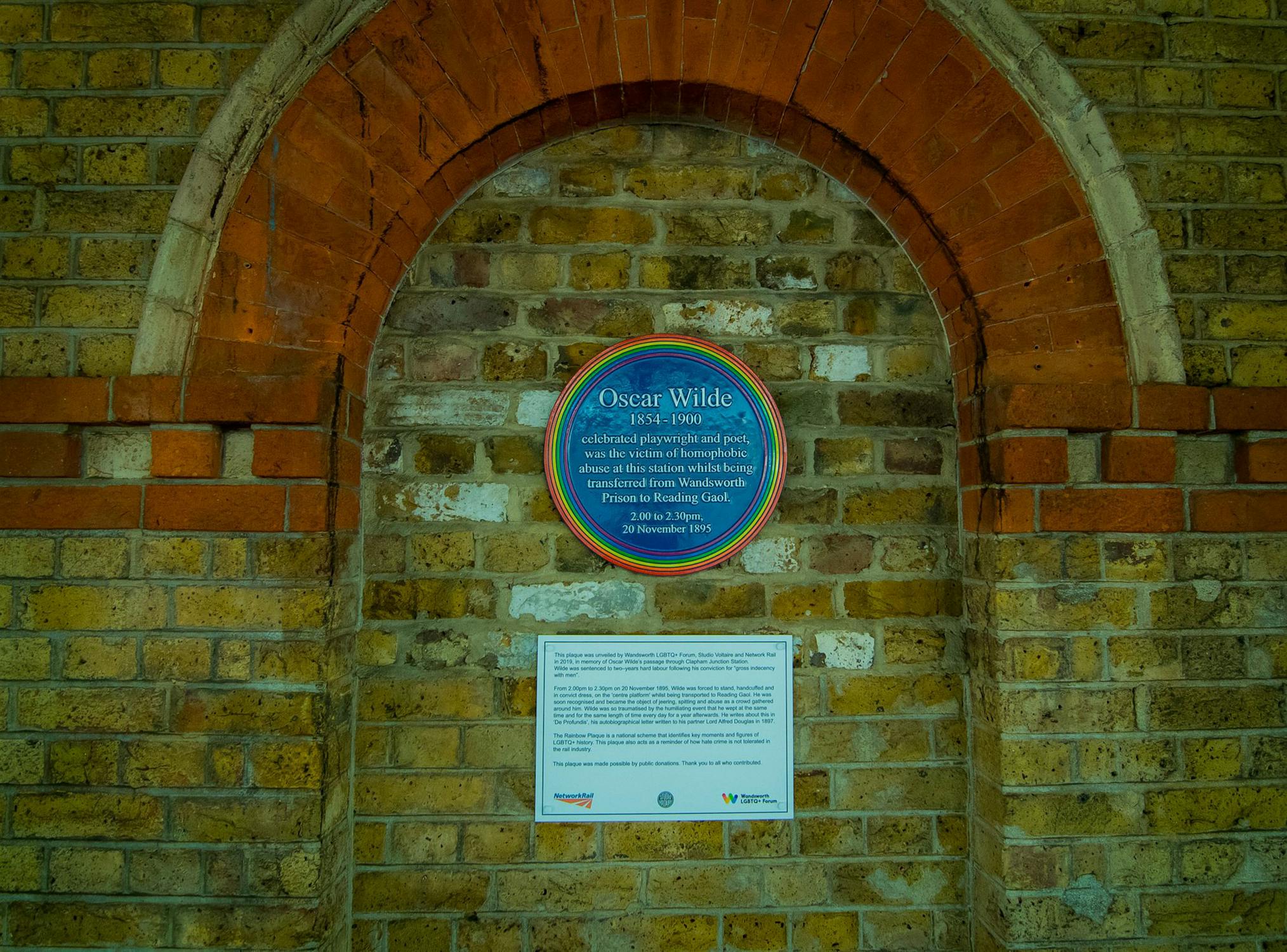 A circular blue plaque with a rainbow edge honouring Oscar Wilde is mounted on a brick wall set within an arched recess. Below the plaque is a rectangular information sign with text.