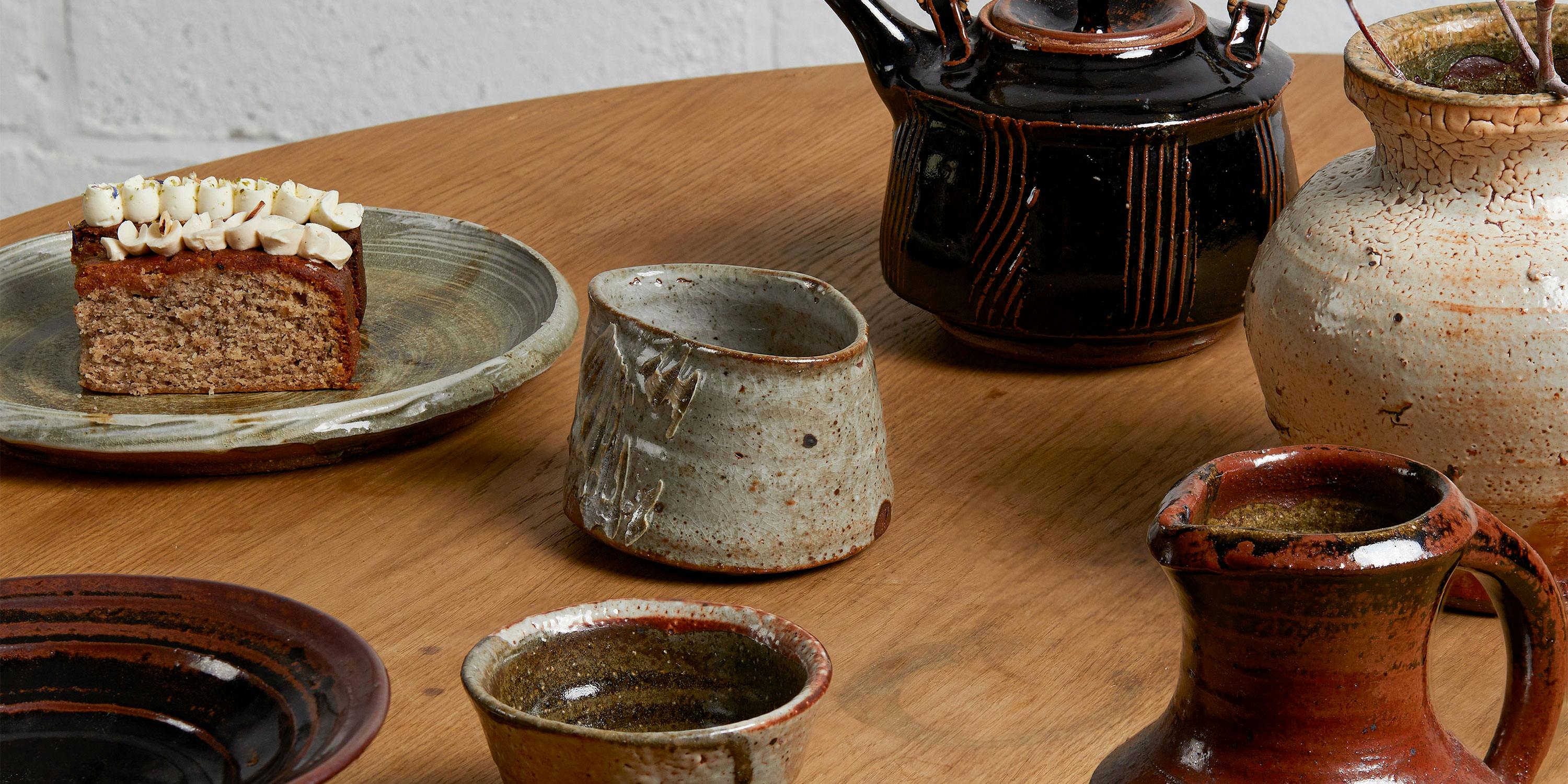 A wooden table set with handmade ceramic cups, pitchers, and plates, including a slice of frosted cake on a ceramic plate. The ceramics vary in earthy tones of brown, cream, and green.