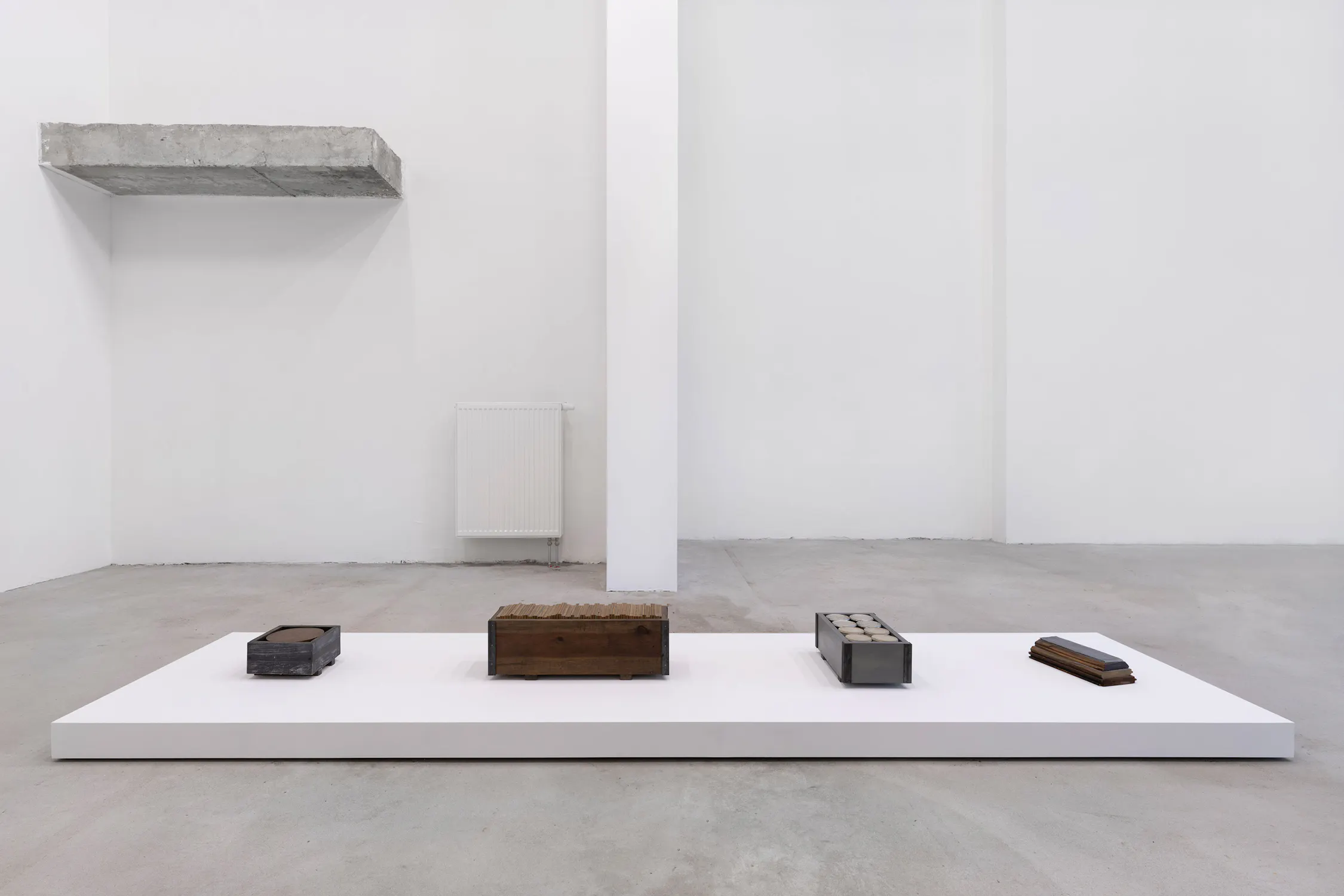Rectangular sculptures by Bill Walton made of metal, concrete, and wood rest on a low white plinth in a minimalist gallery. The background features a white wall, a radiator, and a concrete ledge mounted high on the left side. The floor and walls are plain and greyish.