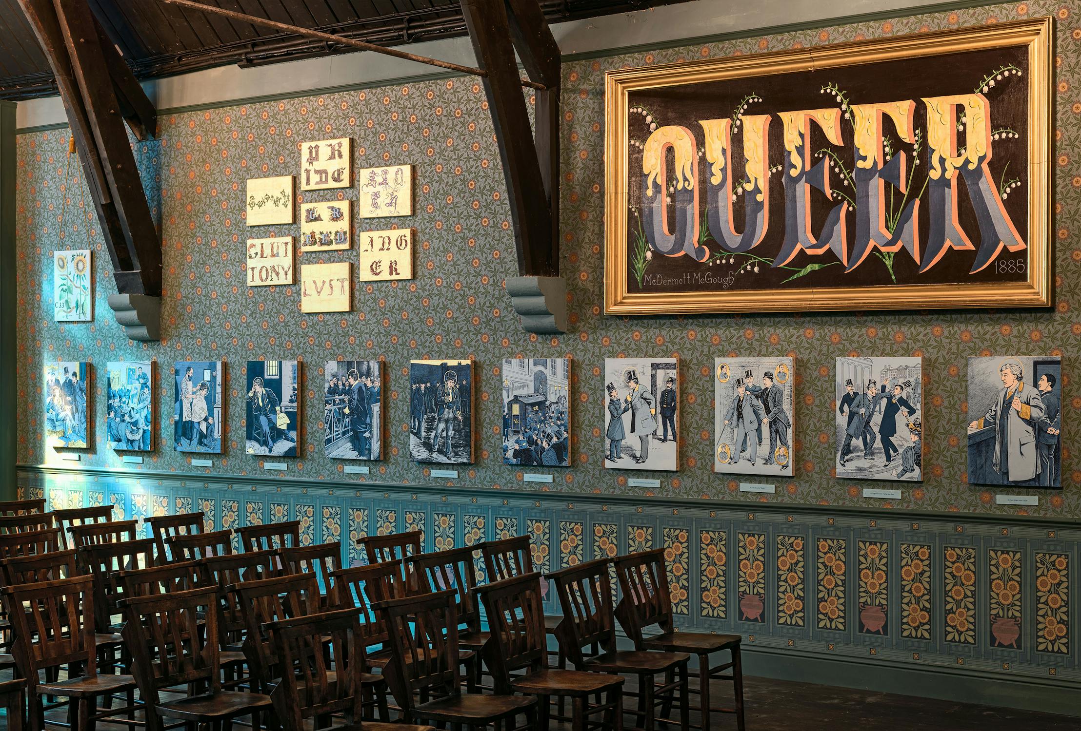 A decorated room with empty wooden chairs faces a wall displaying framed art. Above, a large ornate sign says “QUEER” in bold letters. The patterned wallpaper has floral designs, and the room is lit warmly, highlighting art and historic details.