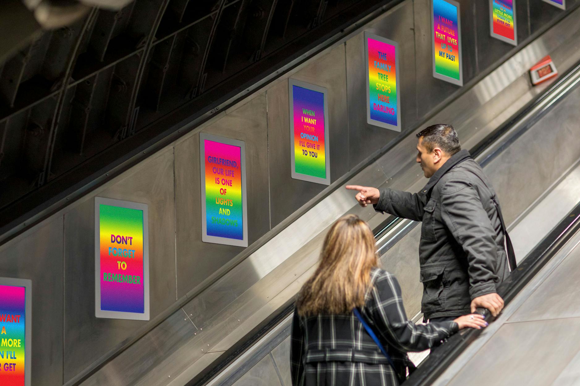 Two people ride an escalator On the London Underground lined with colourful posters by David McDiarmid featuring inspirational quotes written in bold text. One person points at a poster while the other looks up at it.