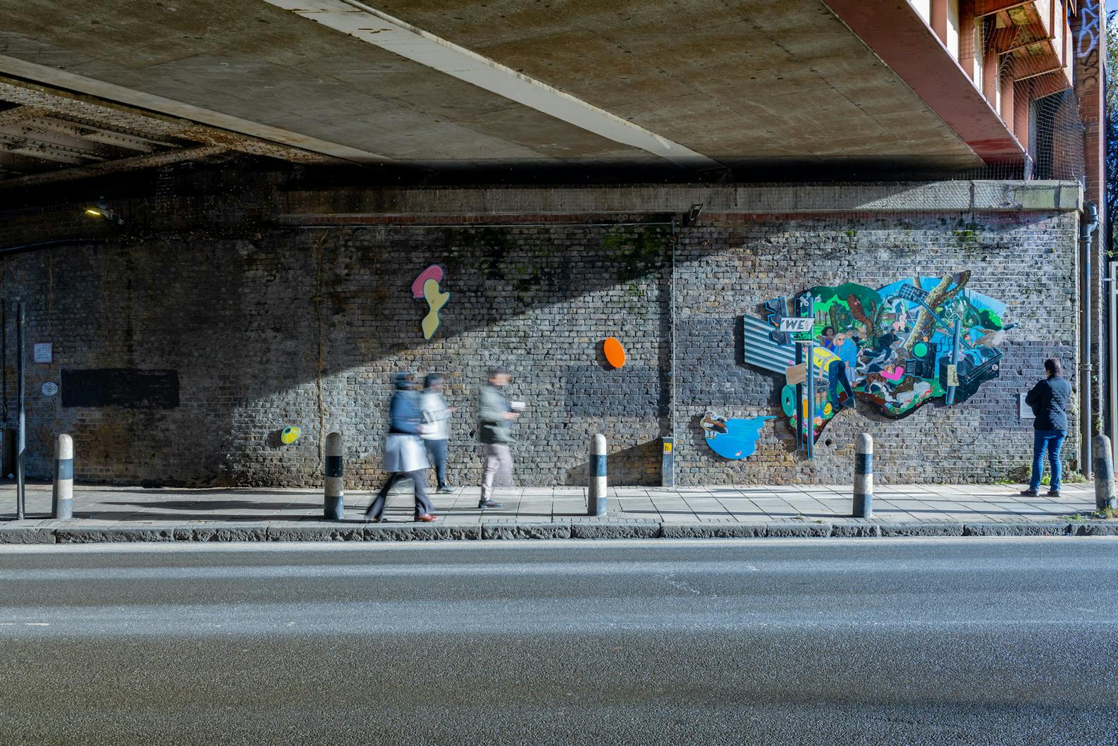 A vibrant and eclectic mural by Flo Brooks is painted on a brick wall. The mural features various elements like a man holding a baby, wildlife such as birds, and urban imagery including street signs and graffiti. There's also a small plaque with text on the wall beside the mural.