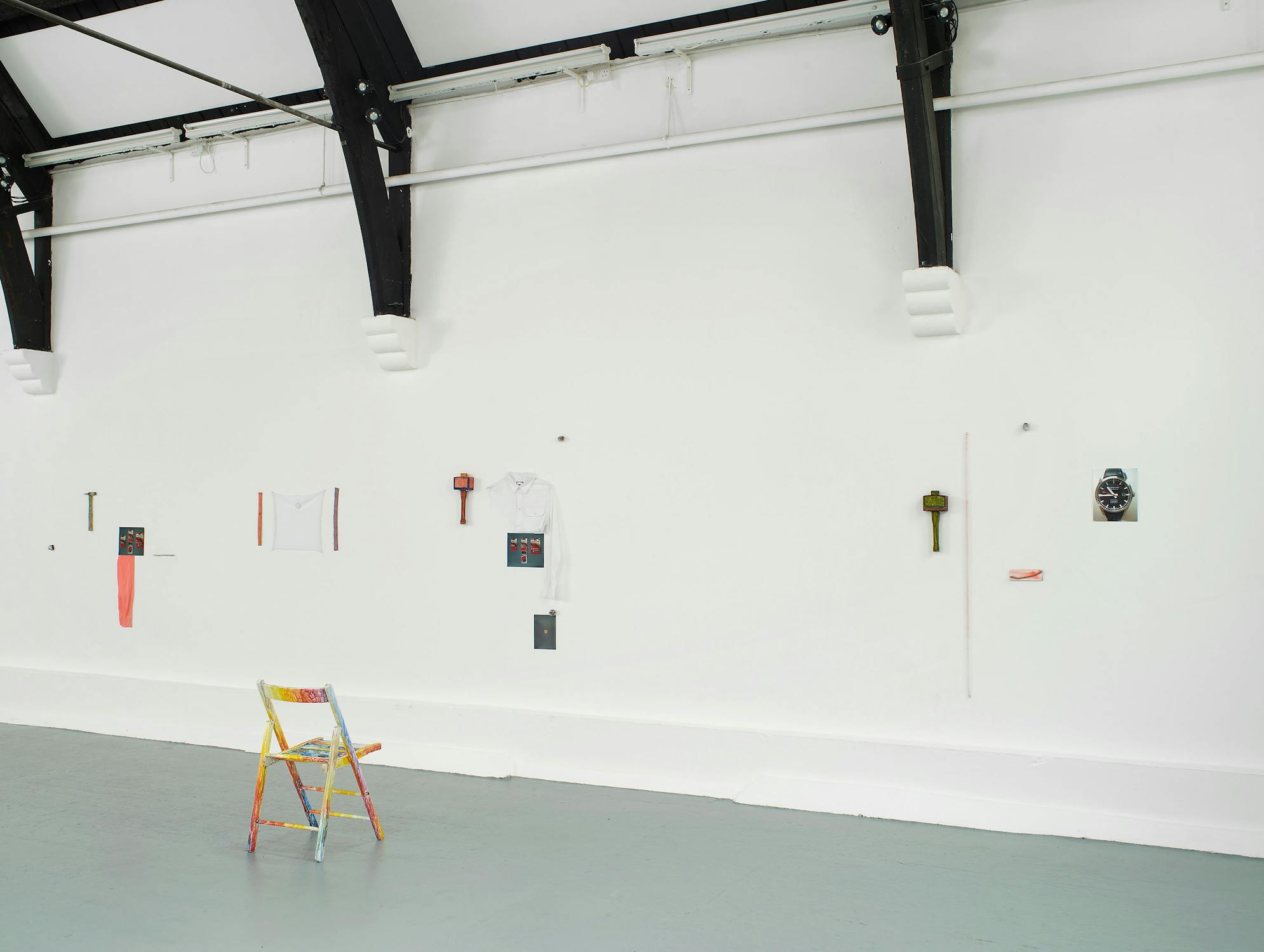 A colourful, painted folding chair faces a white wall with scattered small objects, photos, and art pieces neatly attached. The room features a high ceiling with exposed black beams and soft, even lighting. The overall atmosphere is minimal and modern.