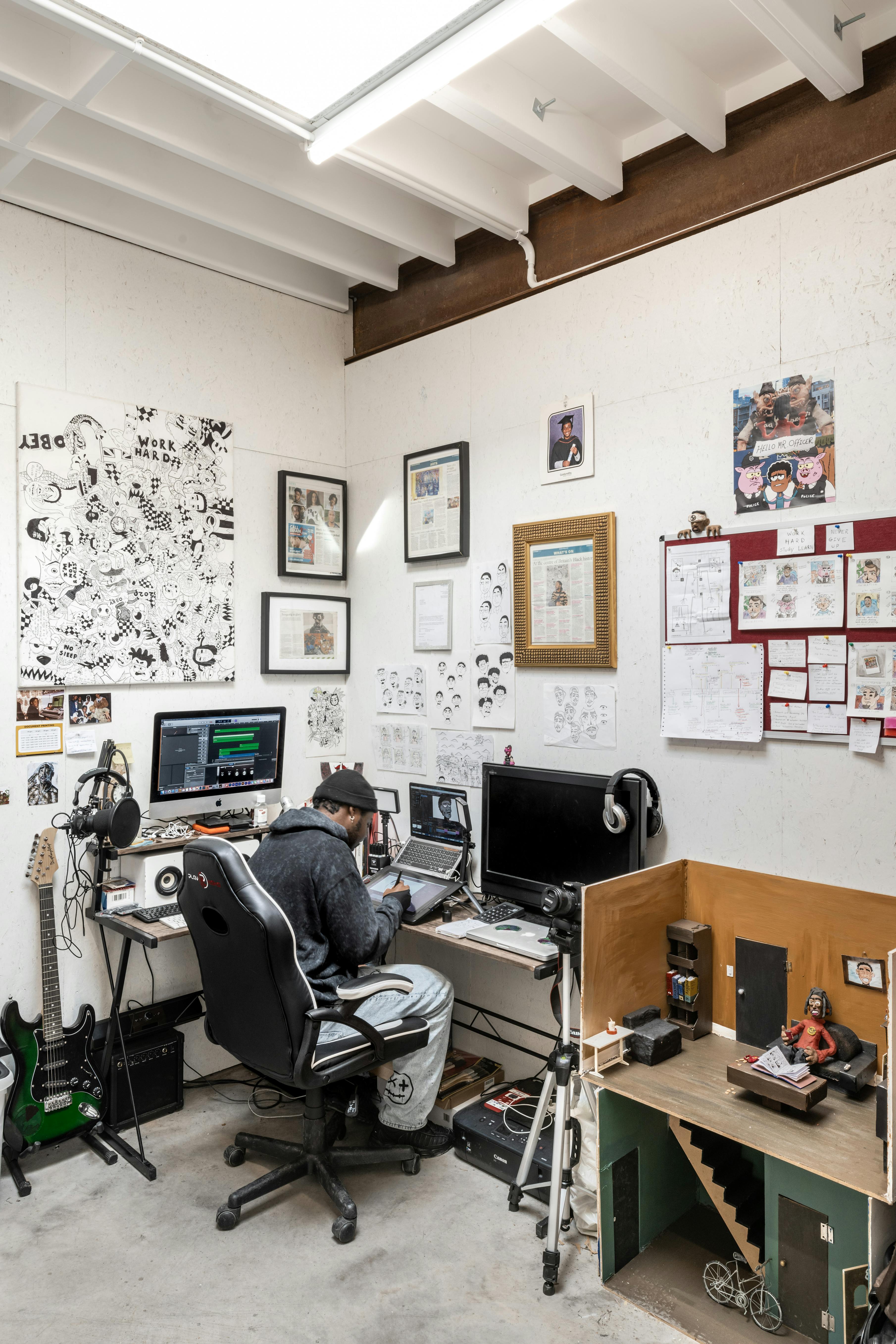 A person in a grey hoodie and cap sits at a cluttered desk with two monitors, audio equipment, and art supplies. The white walls are decorated with sketches, framed art, and photos. A small model house with rooms and tiny furniture sits beside the desk.