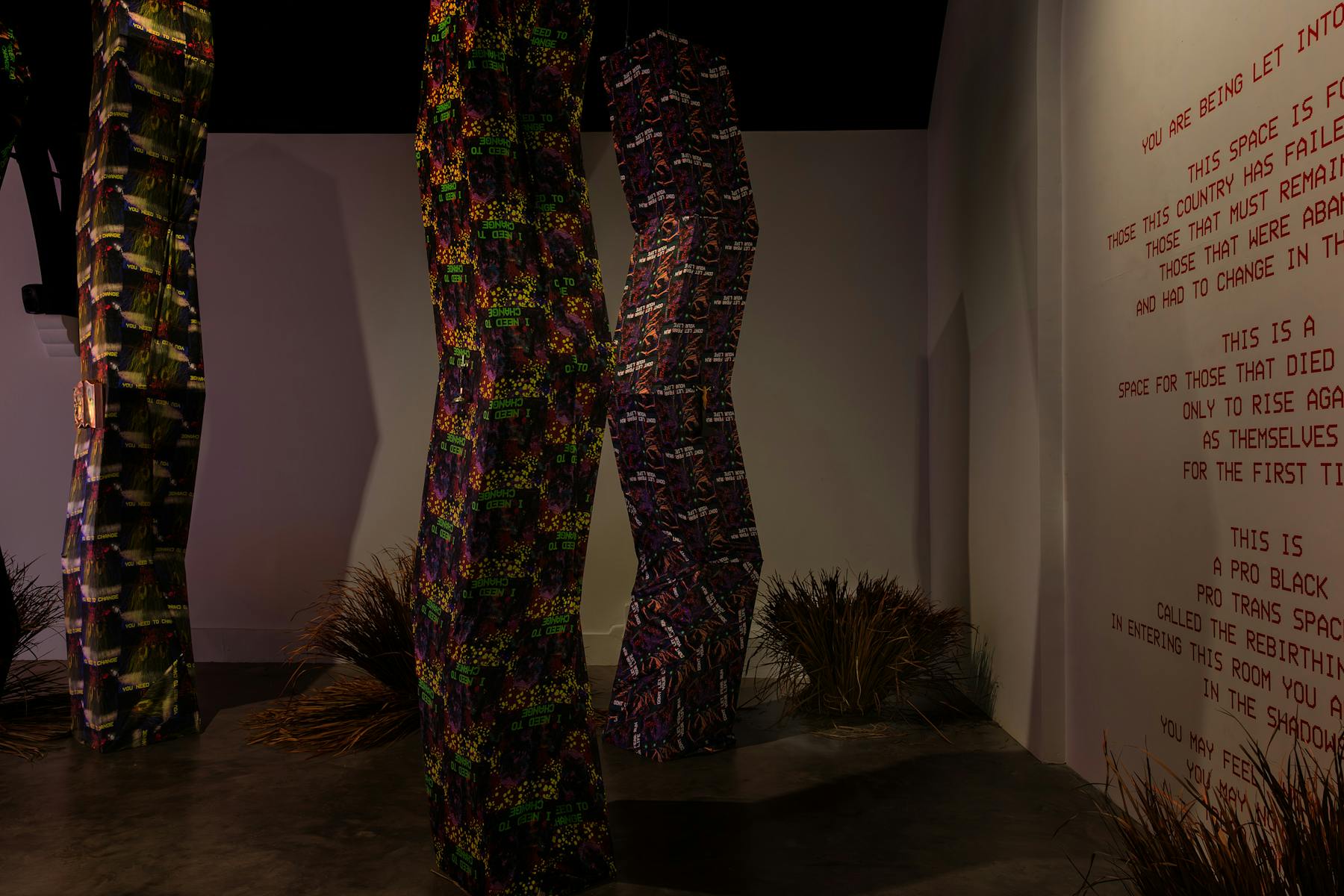 A photograph of a dimly lit room, that is filled with tall angular structures that reach from the floor to the ceiling. The structures are wrapped in vibrantly patterned fabrics. a wall to the right has a set of instructions in red letters.