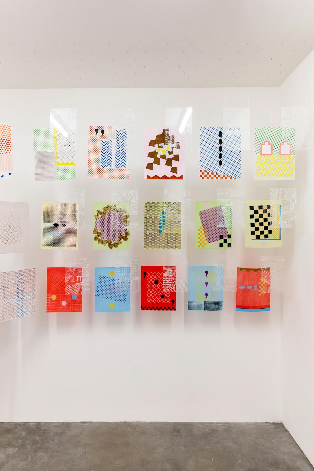 A white gallery wall displays a grid of colourful abstract artworks by Bamidele Awoyemi and Adia Wahid. Each piece features various patterns, shapes, and colors, including reds, greens, blues, and yellows. The floor is concrete, and the room is well-lit.