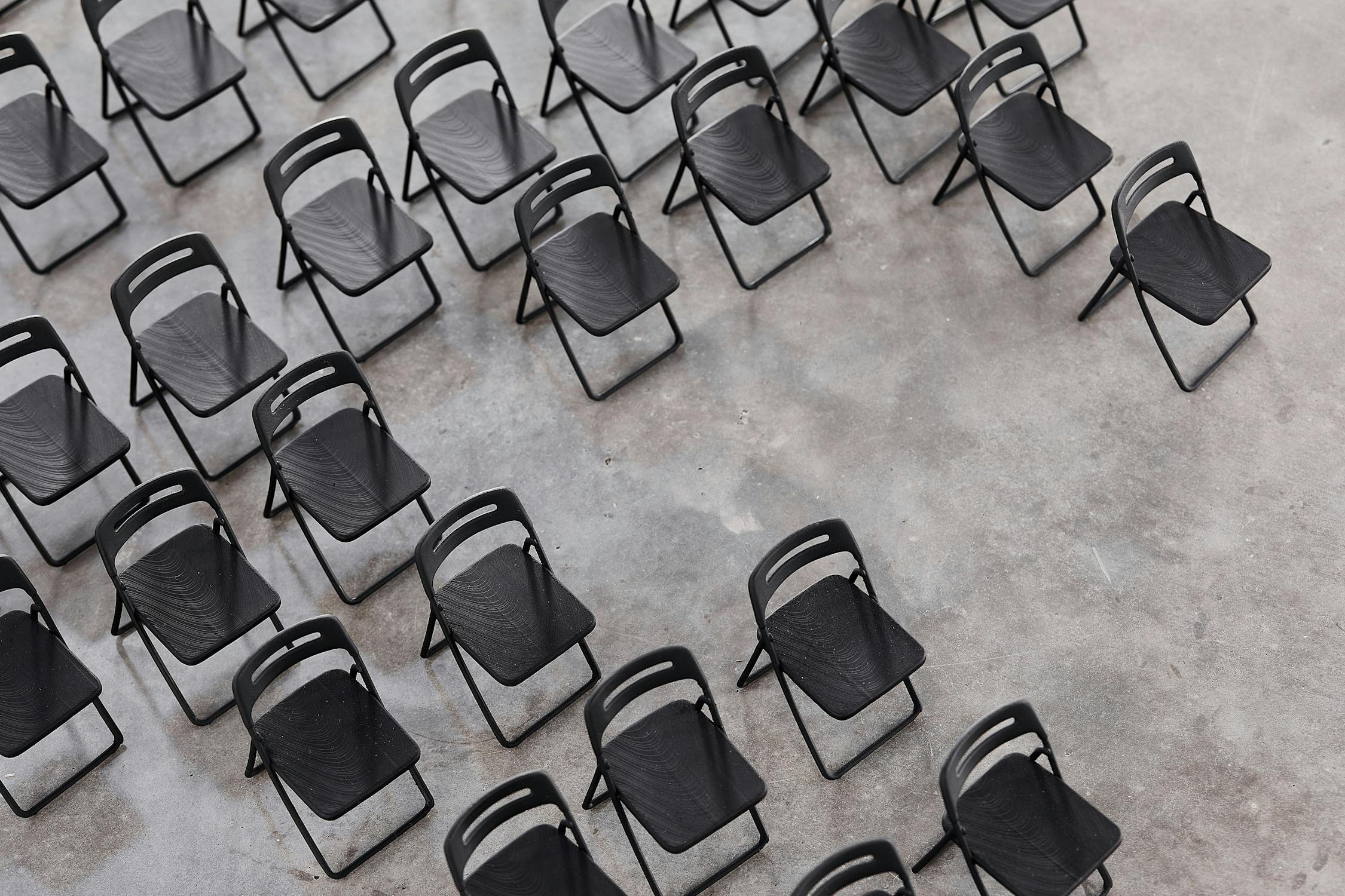 Rows of empty 3D printed small black folding chairs are arranged at an angle on a grey concrete floor, leaving space on the right side of the image.