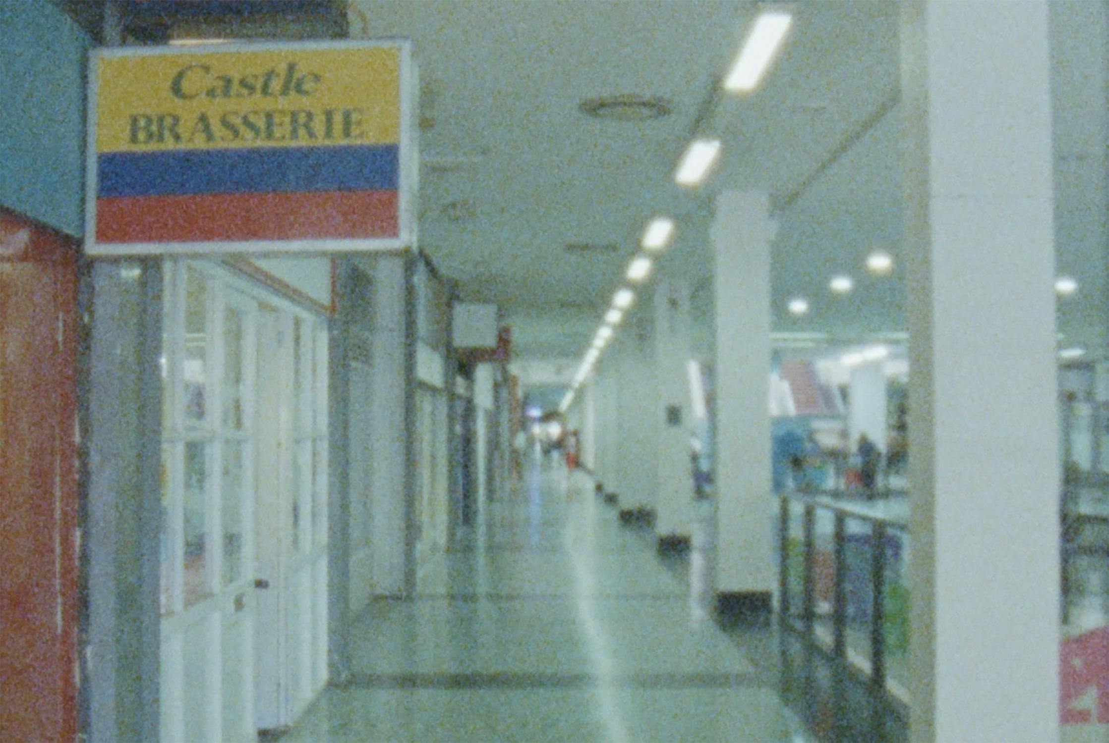 A vintage photo of a shopping mall corridor with fluorescent lighting and a retro sign for "Castle Brasserie" on the left. The hallway features tiled flooring and leads to various shops visible in the distance.