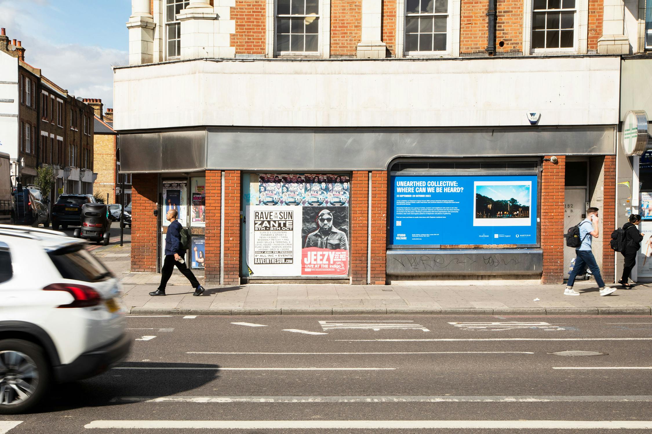 A city street with pedestrians walking by a building displaying various posters. A car drives past on the road in the foreground. The building has brick and light-colored walls, and the sky is partly cloudy.