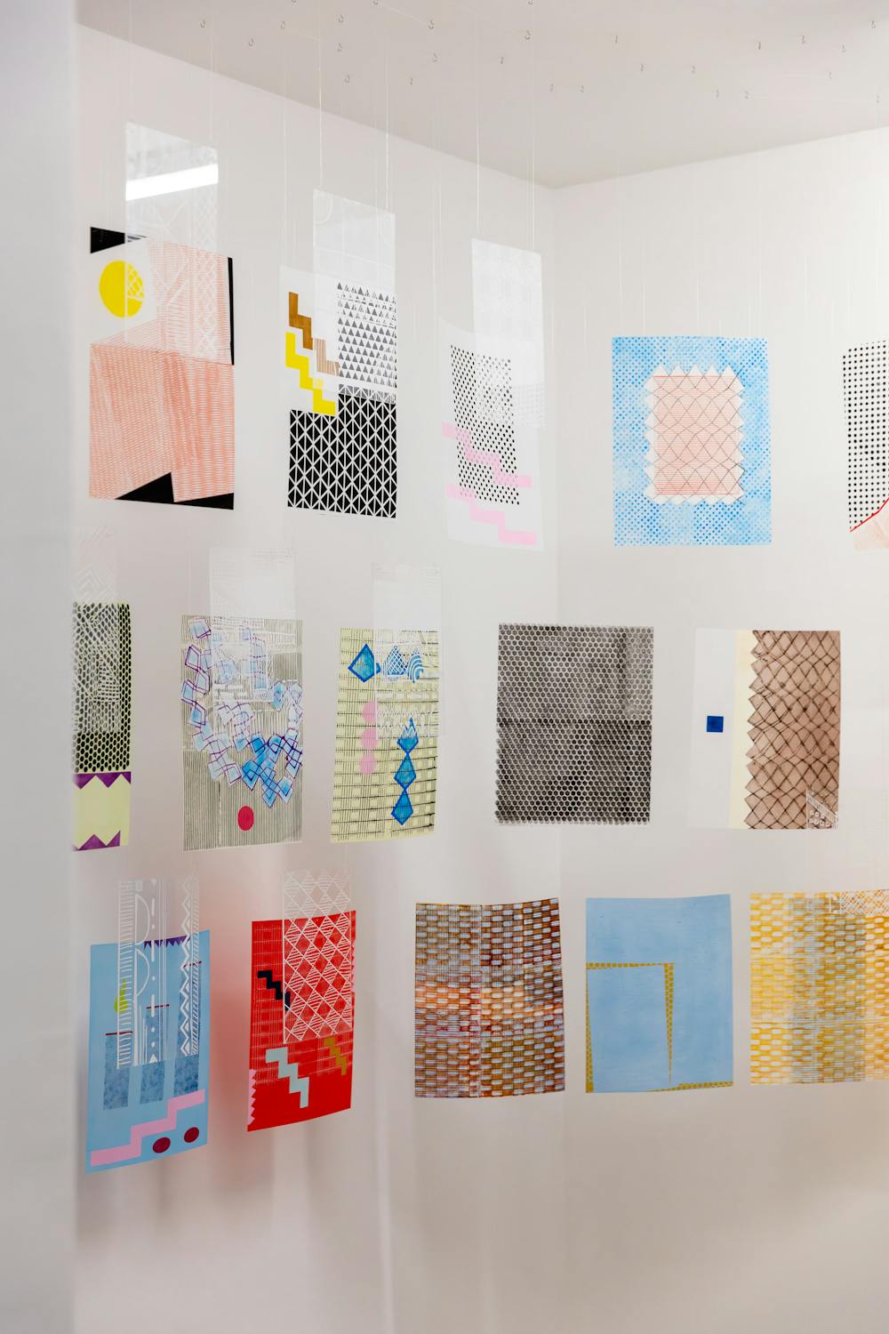 A collection of colourful abstract art pieces by Baimdele Awoyemi and Adia Wahid hanging in a gallery space. The artworks feature various patterns and textures, including geometric shapes and grids in shades of pink, blue, yellow, and more. They are suspended from the ceiling.