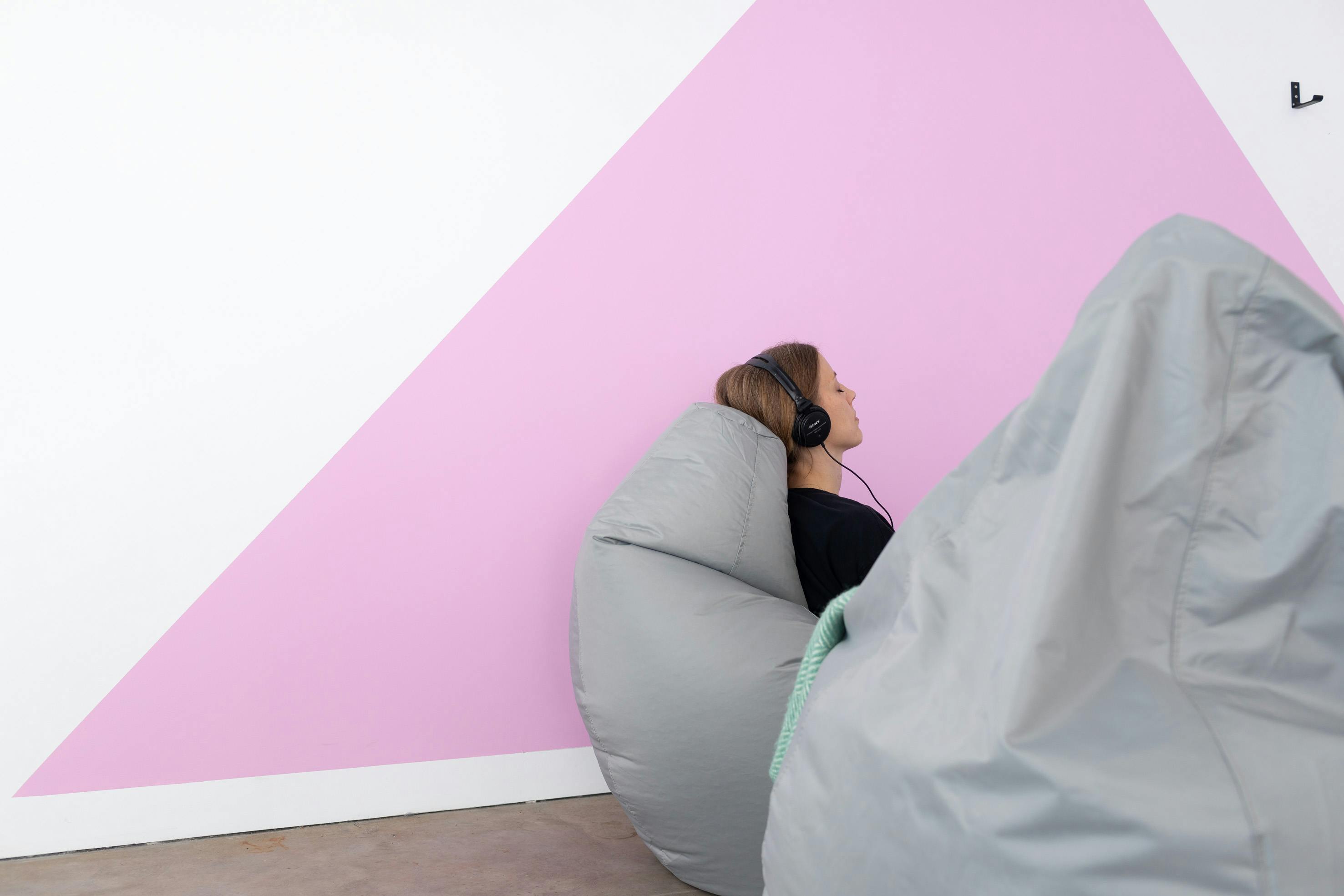 a photograph of a woman wearing headphones sitting on a large grey bean bag. The wall in the background is painted white with a large bright pink triangle.