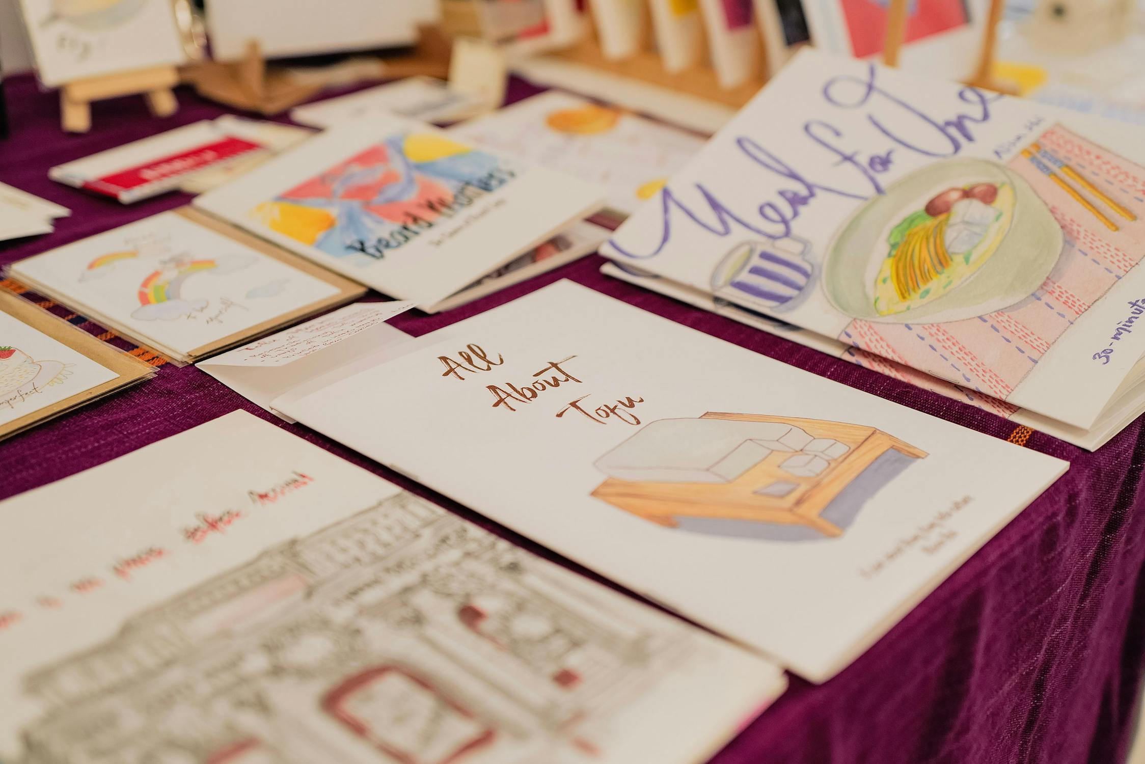 A close-up of illustrated zines and booklets displayed on a purple surface, including one titled "All About Tofu" featuring a drawing of tofu on a table.