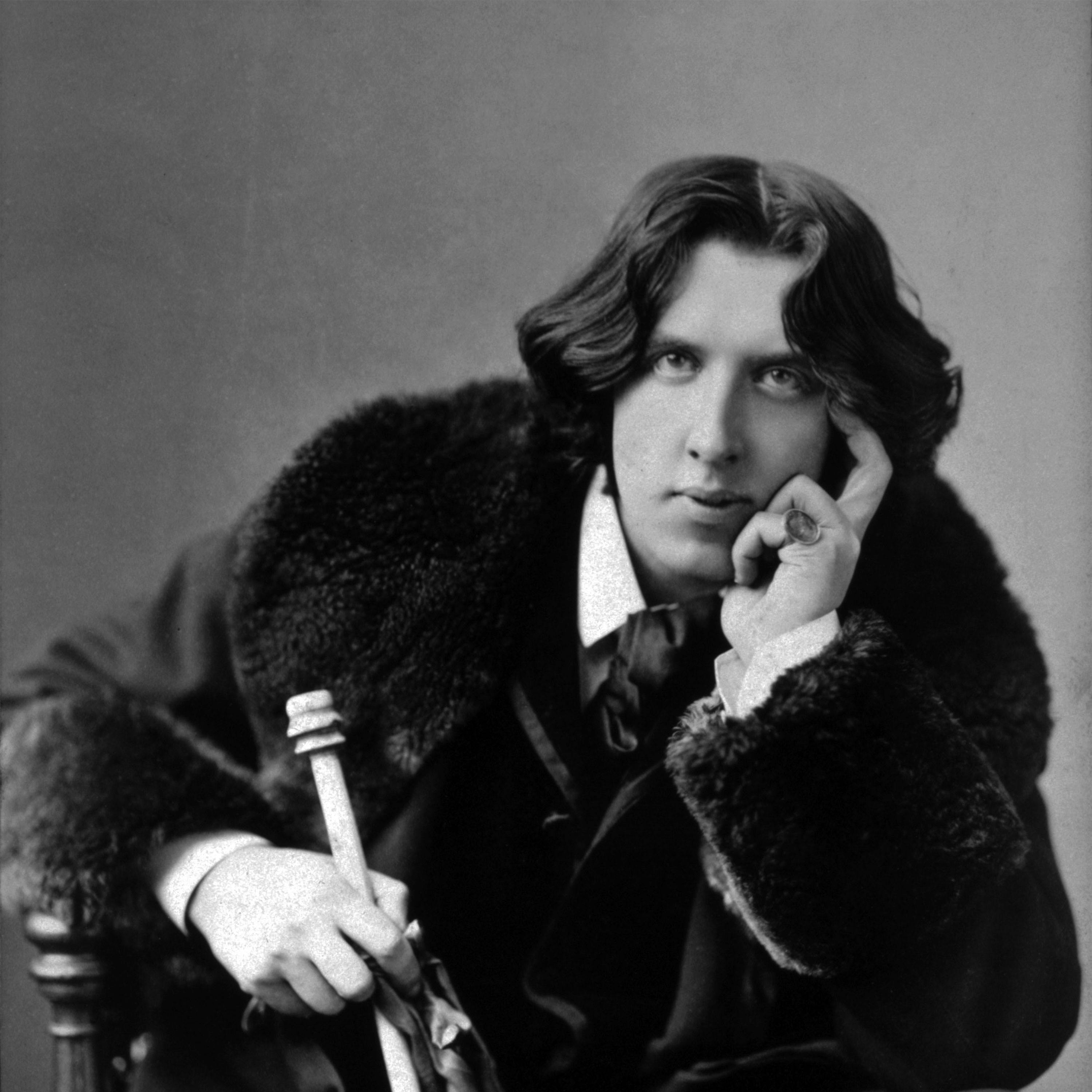 A black-and-white portrait of Oscar Wilde, with wavy, shoulder-length hair, wearing a fur-collared coat. Seated, he rests his head on one hand and holds a cane in the other, his expression thoughtful.