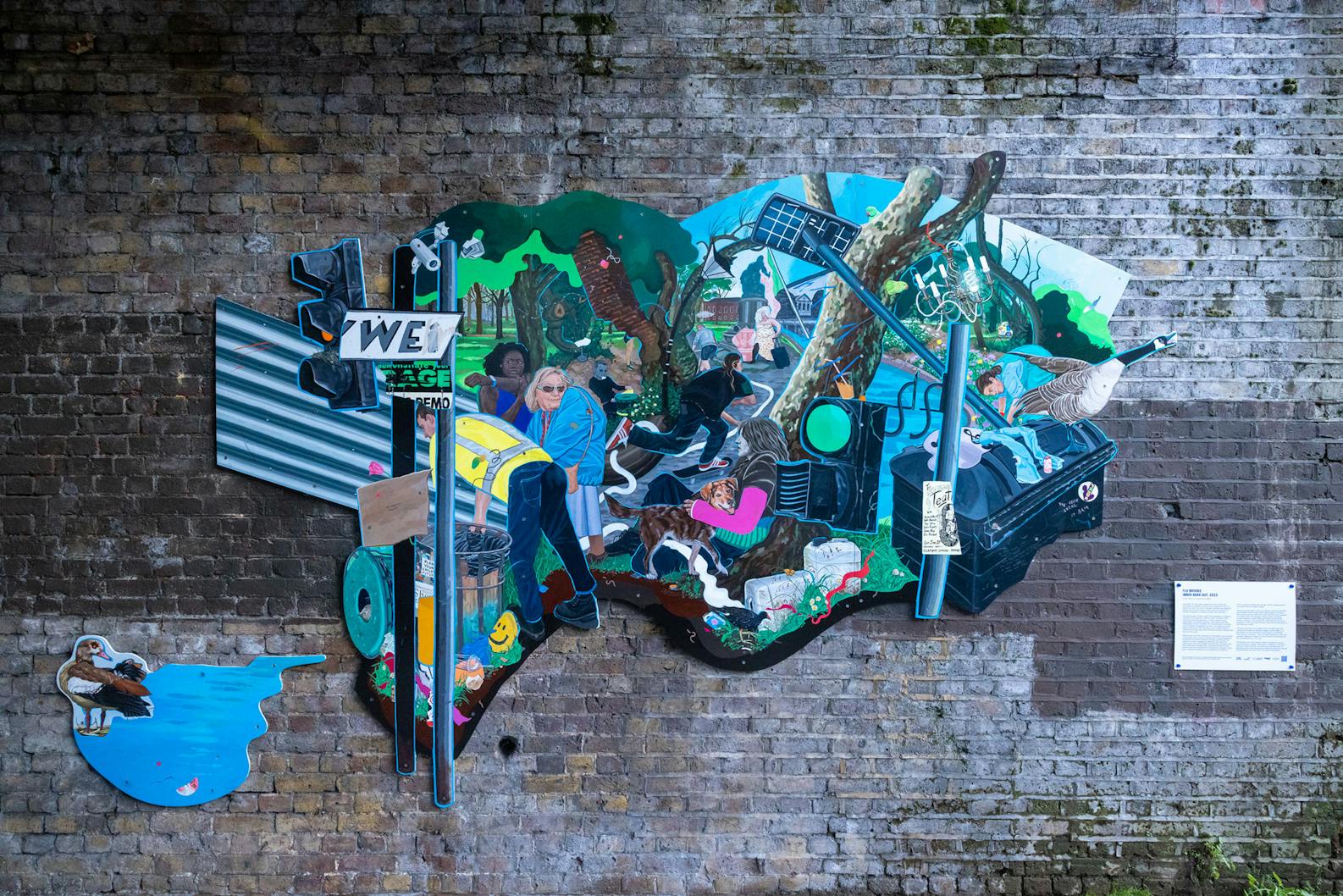 A vibrant and eclectic mural by Flo Brooks is painted on a brick wall. The mural features various elements like a man holding a baby, wildlife such as birds, and urban imagery including street signs and graffiti. There's also a small plaque with text on the wall beside the mural.