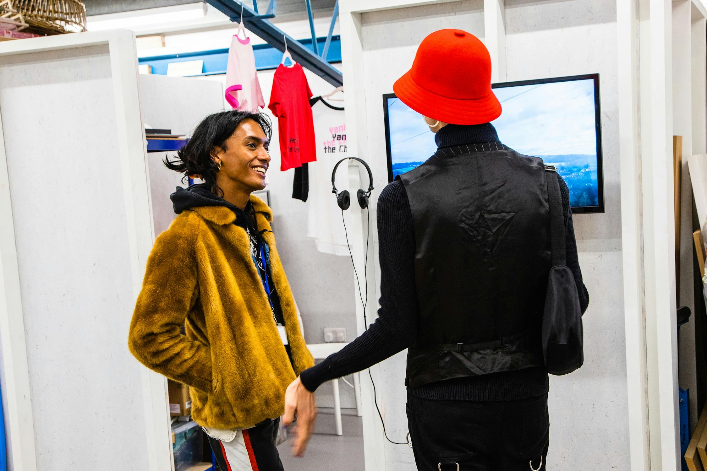 A person wearing a brown fur coat and blue lanyard smiles while talking to someone in a red hat and black vest inside a modern clothing store with T-shirts and headphones on display.