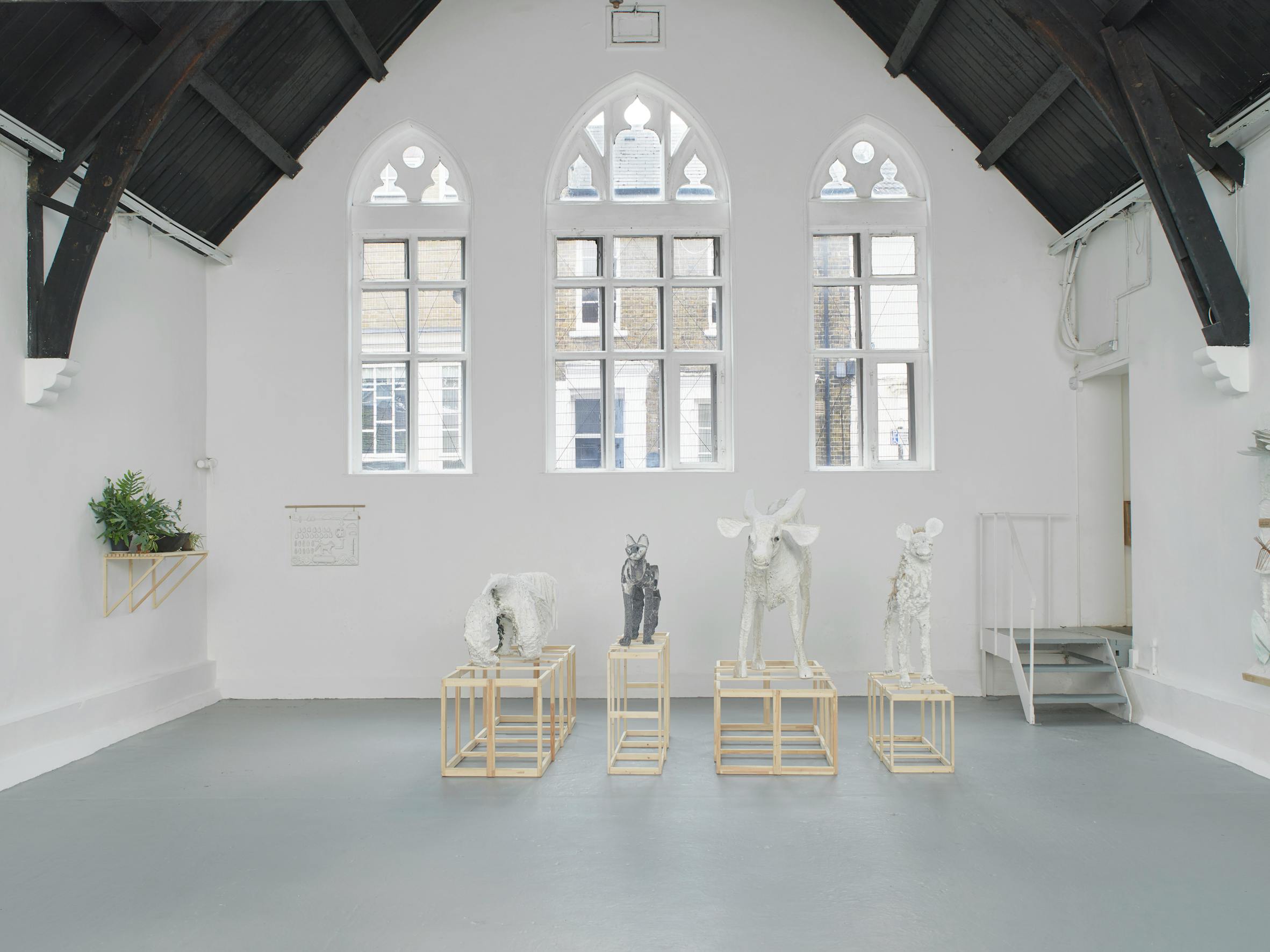 A bright gallery with vaulted black ceilings and arched windows displays four abstract animal sculptures on wooden frames and a large textured white artwork mounted on a wall. The space is minimal, with light grey floors and white walls.