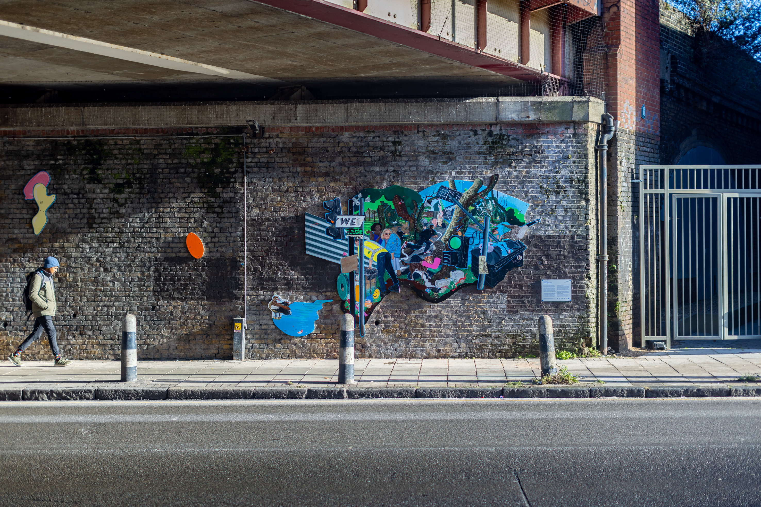 A vibrant and eclectic mural by Flo Brooks is painted on a brick wall. The mural features various elements like a man holding a baby, wildlife such as birds, and urban imagery including street signs and graffiti. There's also a small plaque with text on the wall beside the mural.