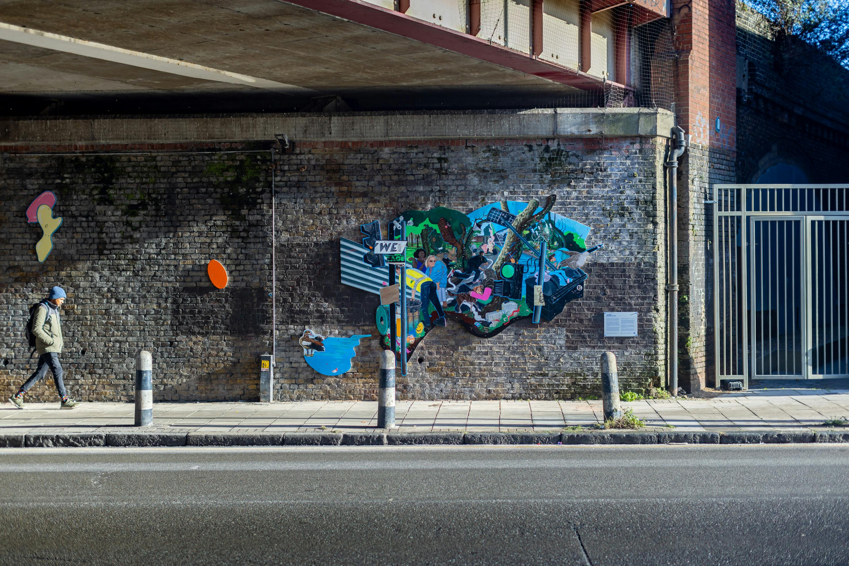 A vibrant and eclectic mural by Flo Brooks is painted on a brick wall. The mural features various elements like a man holding a baby, wildlife such as birds, and urban imagery including street signs and graffiti. There's also a small plaque with text on the wall beside the mural.