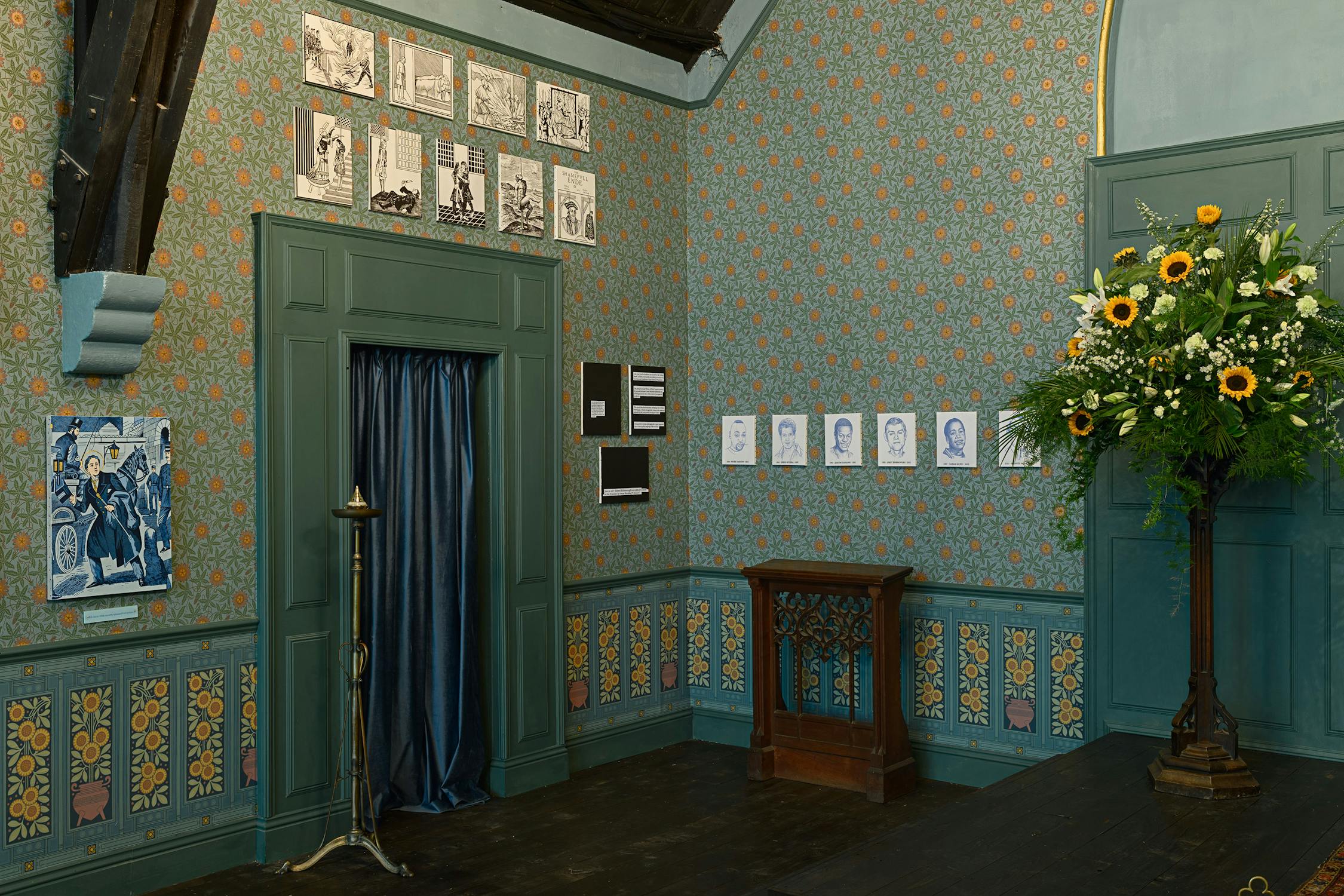 A richly decorated room with blue-green floral wallpaper and panelling. Framed sketches hang on the walls, with a blue curtain at centre. A large bouquet with sunflowers sits in the corner. A wooden stand and a tall candlestick are also visible.