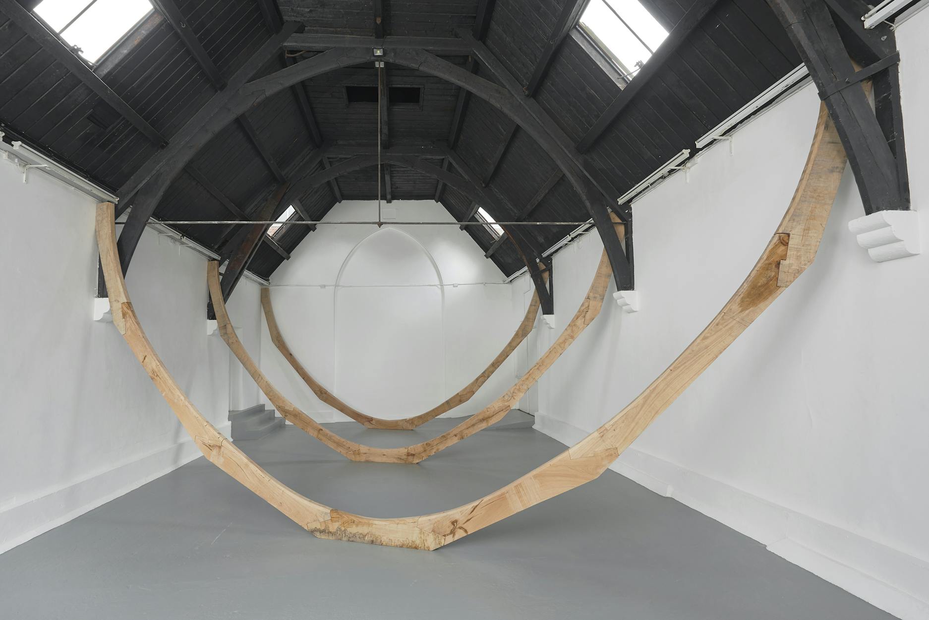 Large wooden arcs are suspended from the ceiling of a white, vaulted gallery space with dark beams and high windows, forming sweeping curves across the room.