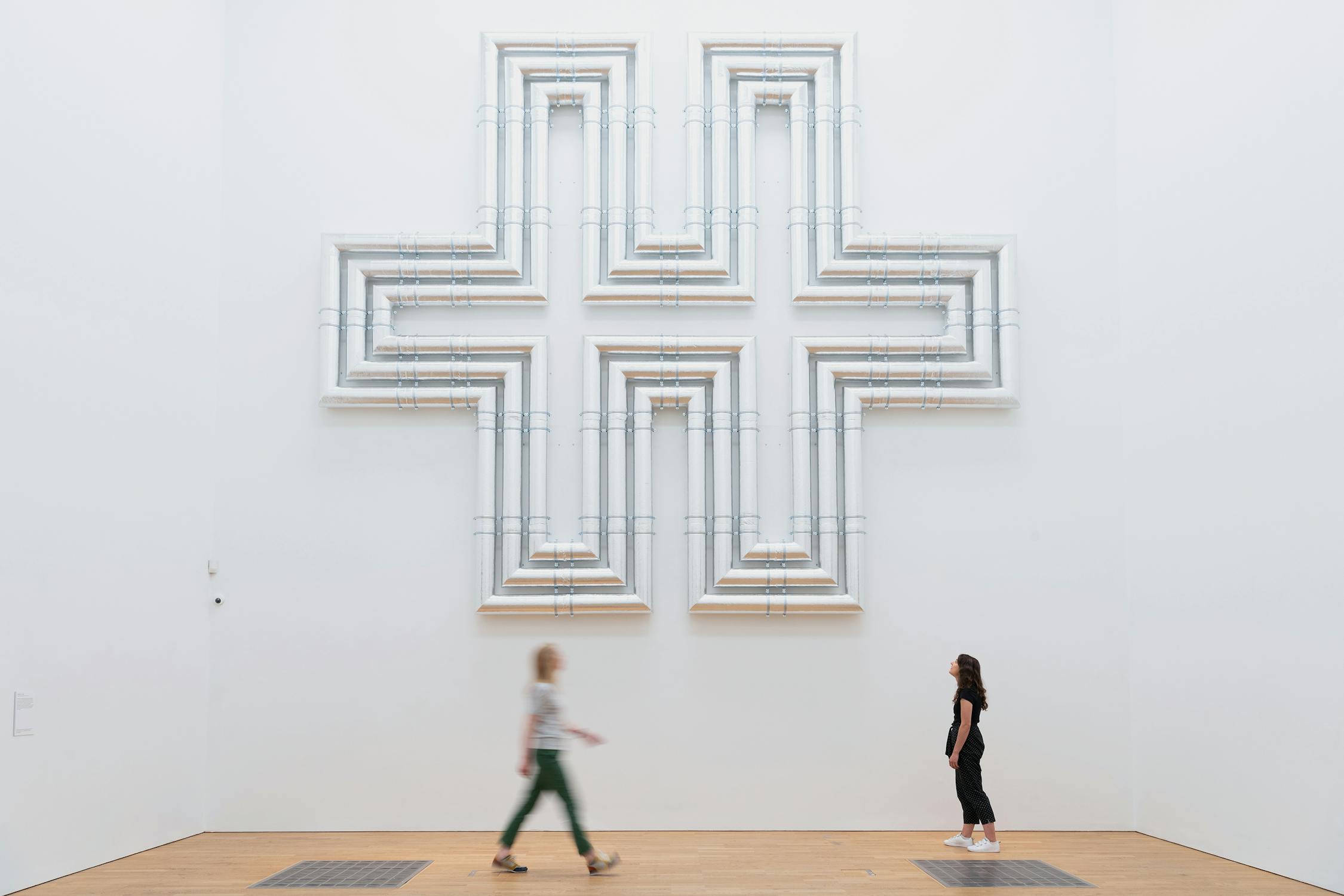 Two people walk past a large, geometric wall sculpture by Lubna Chowdhary. The sculpture is in the shape of a cross, composed of layered white and metallic lines. The artwork is displayed against a plain white wall in a spacious room.