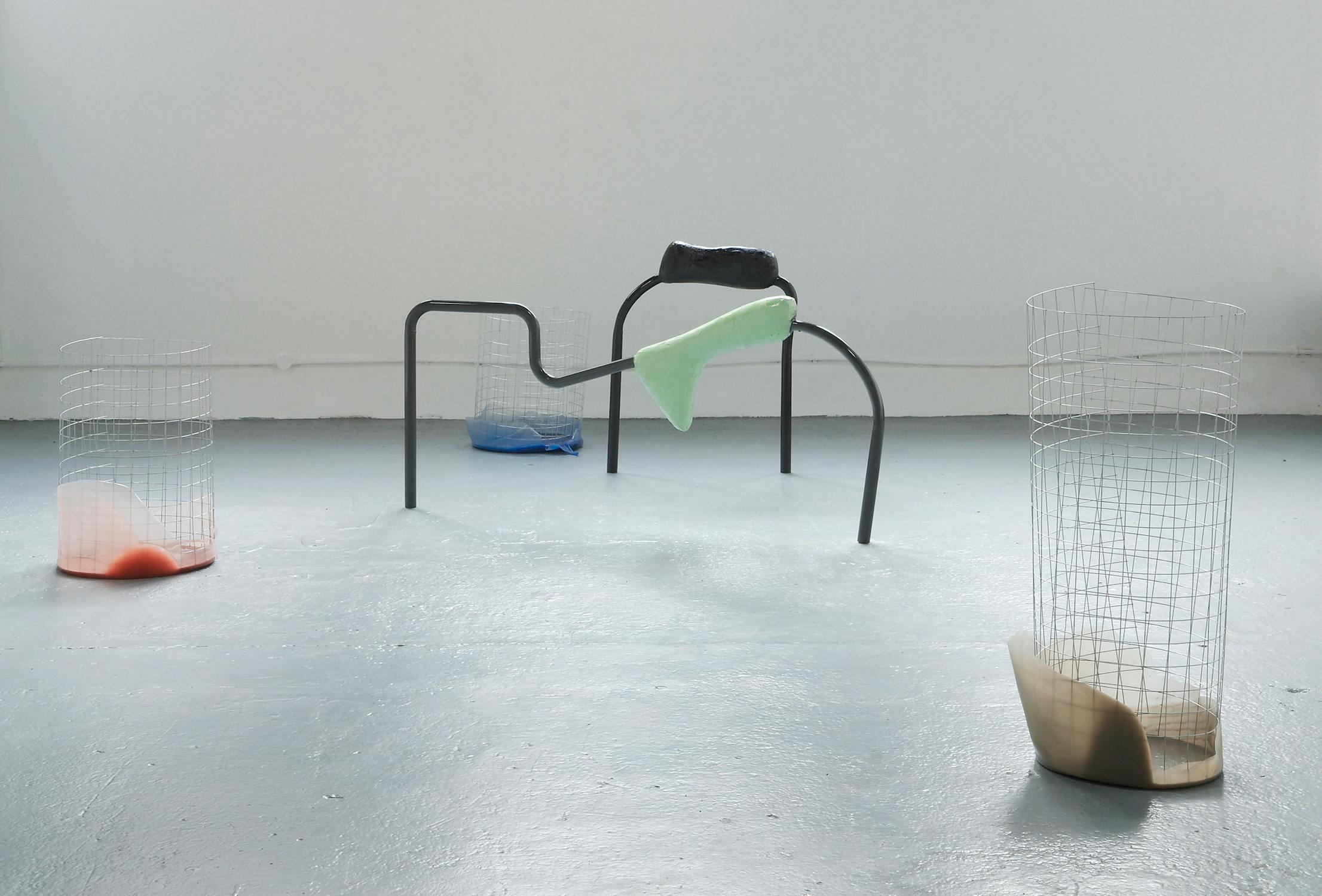 A minimalist art installation featuring abstract metal and wire structures on a smooth grey floor. Each structure contains colourful latex-like material in colours like orange, blue, green, and beige. The scene is in a well-lit, white-walled space.