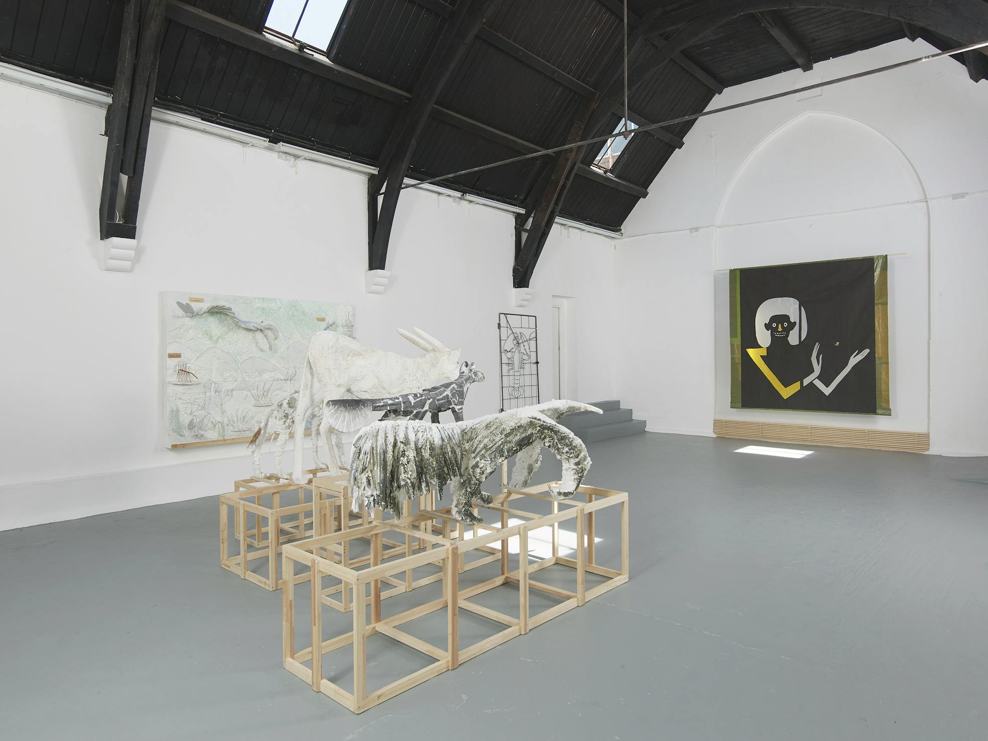 A bright art gallery with high black ceilings displays abstract sculptures on wooden frames, a textured painting on the left wall, and a large tapestry featuring a stylised figure with a skull-like face and raised hand on the right wall.