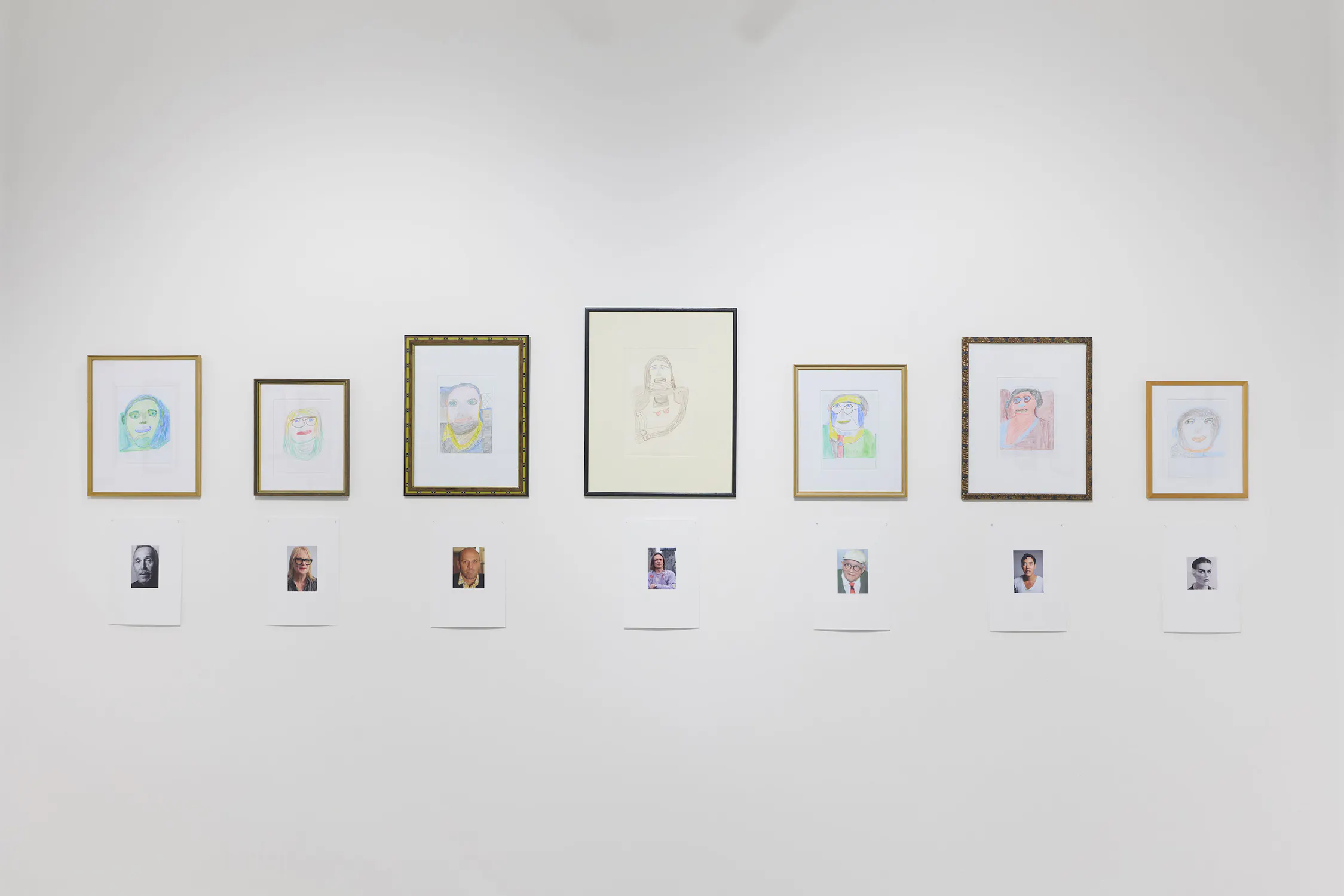 Seven framed drawings of faces are hung on a white wall, each with a small colour photo and text beneath. The portraits vary in style; all are simple and colourful. The arrangement is orderly, creating a gallery display.