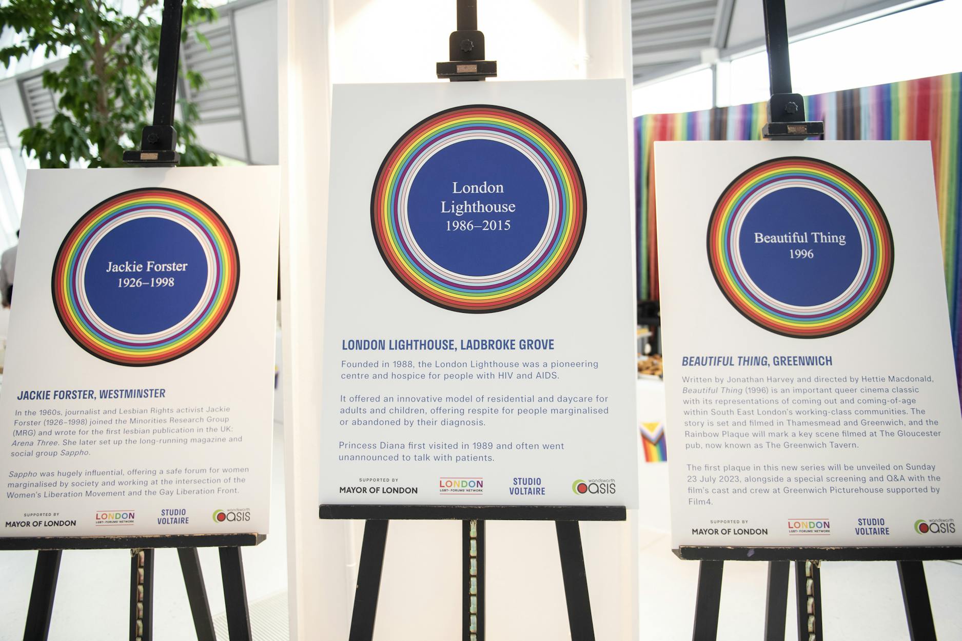 Three display boards with rainbow circle designs, each honouring LGBT history: Jackie Forster (1926–1998), London Lighthouse (1986–2015), and Beautiful Thing (1996), with brief descriptions beneath each title.