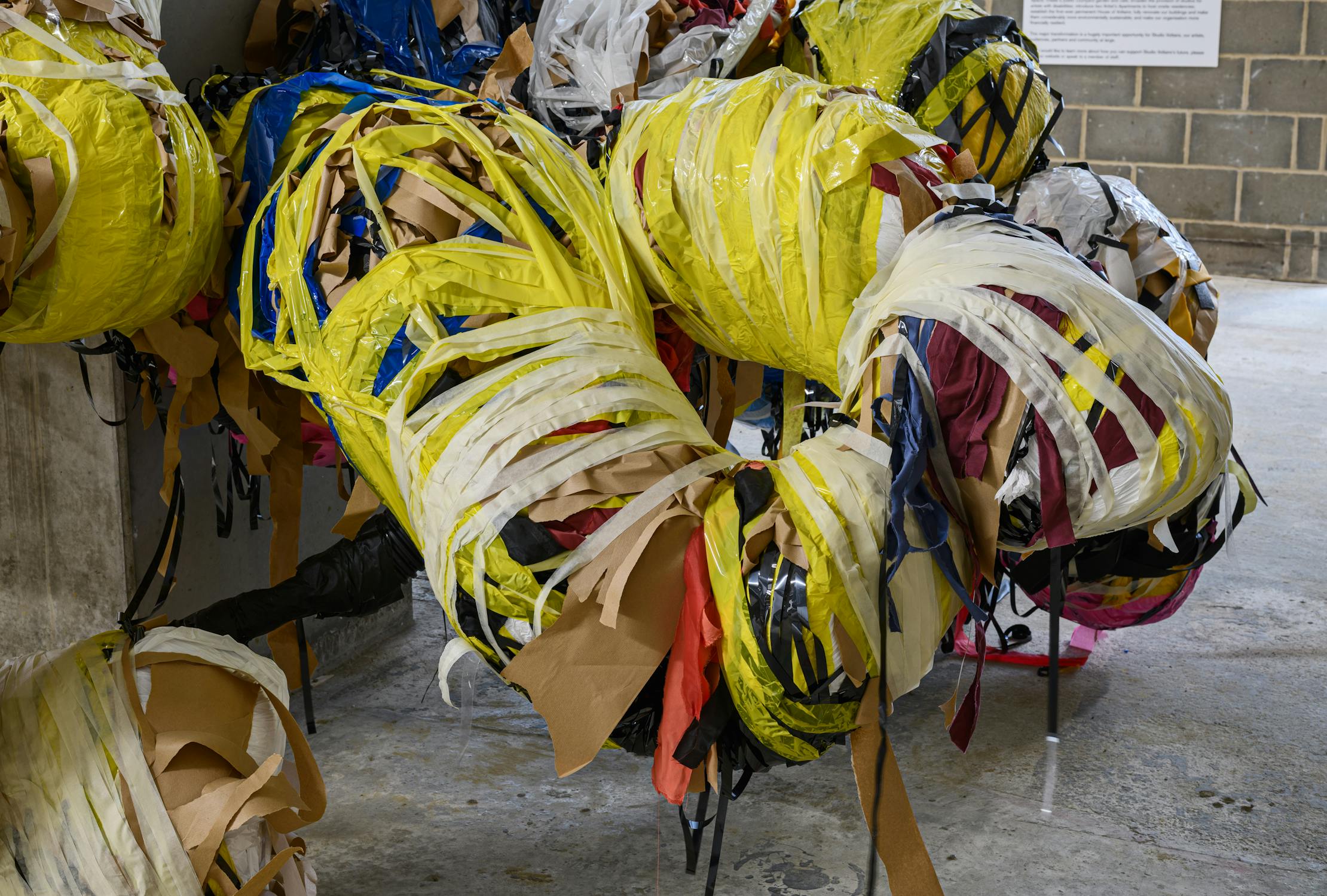 Large abstract sculpture made of tightly wrapped, overlapping strips of yellow, white, and brown plastic and paper. The structure is irregular and bulbous, emerging from a concrete wall in an industrial space with a brick wall and sign in the background.