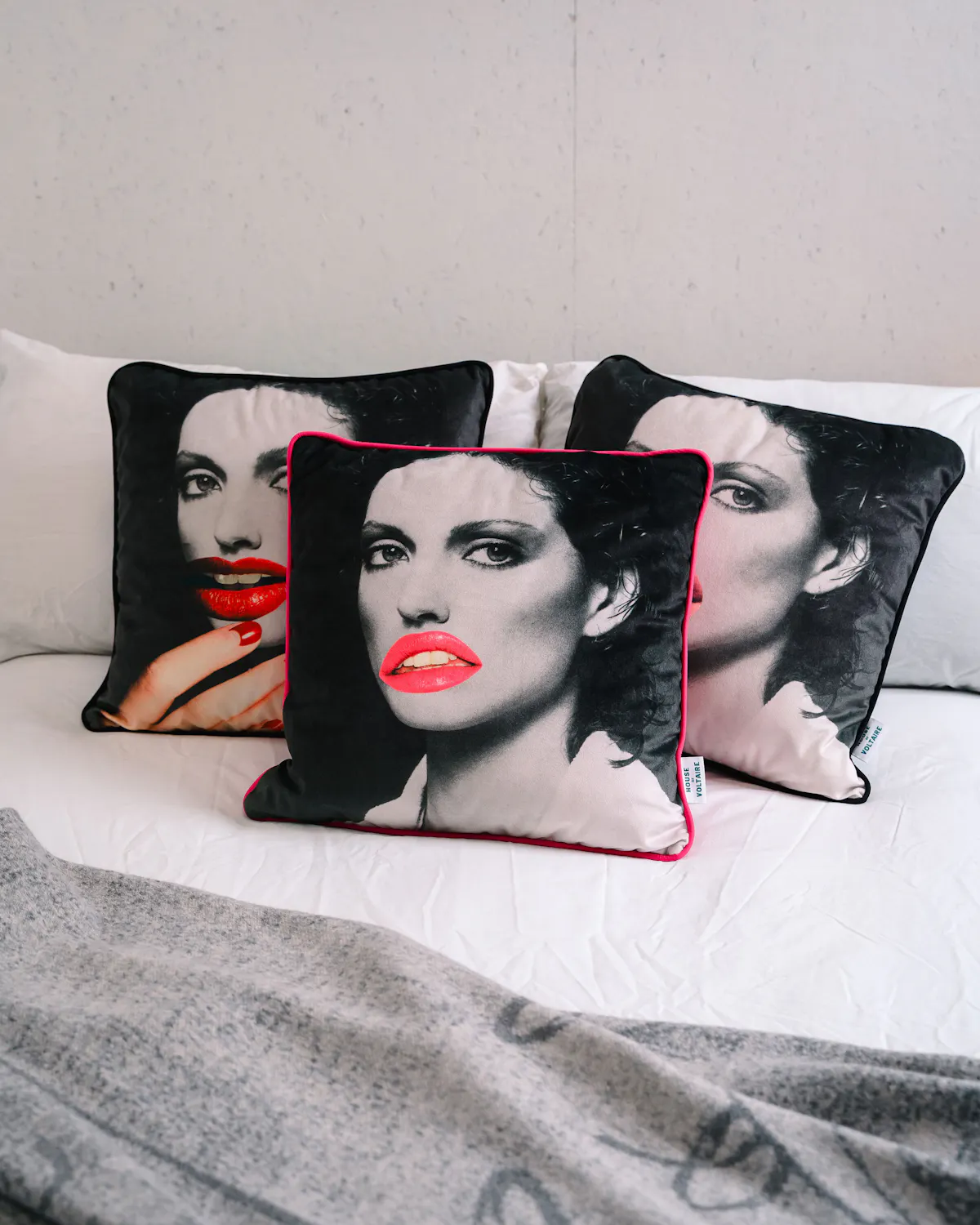 Three square Linder Sterling-designed cushions with a black-and-white face print and bright red lips are arranged on a white bed. A grey blanket is draped over the bed’s lower edge. The background wall is plain.