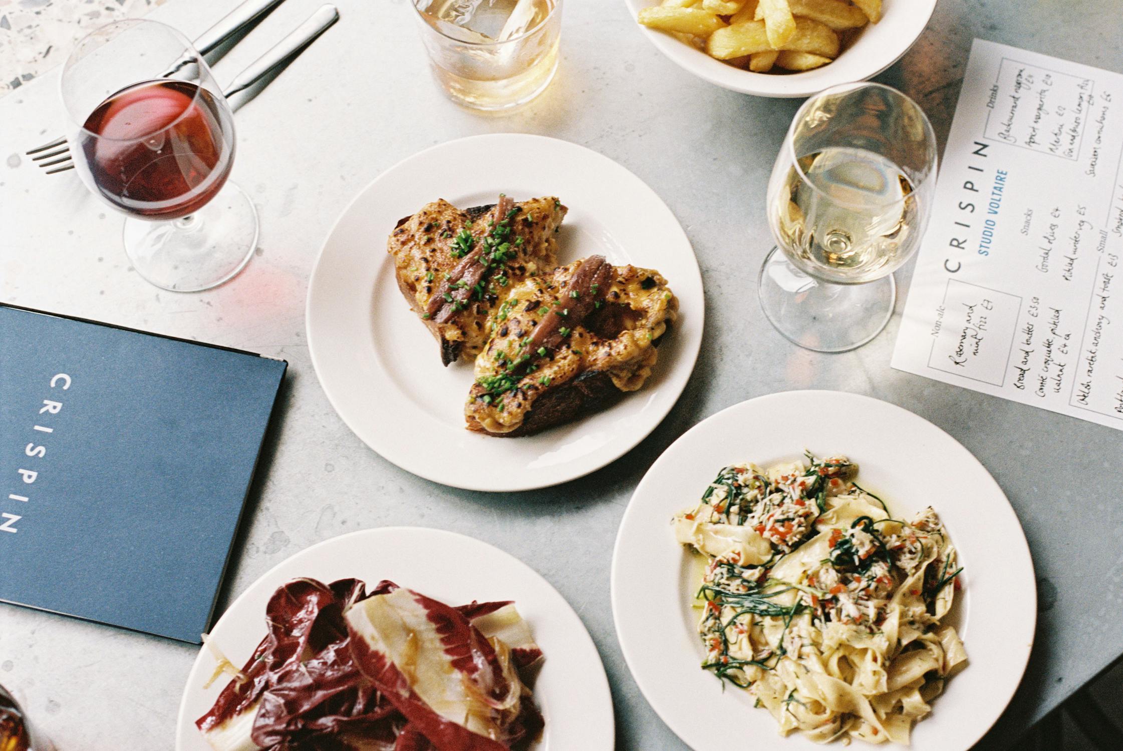 A photograph of spread of freshly prepared restuarat food on white plates, including a creamy pasta dish, dressed leaves, and glasses or red and white wine.