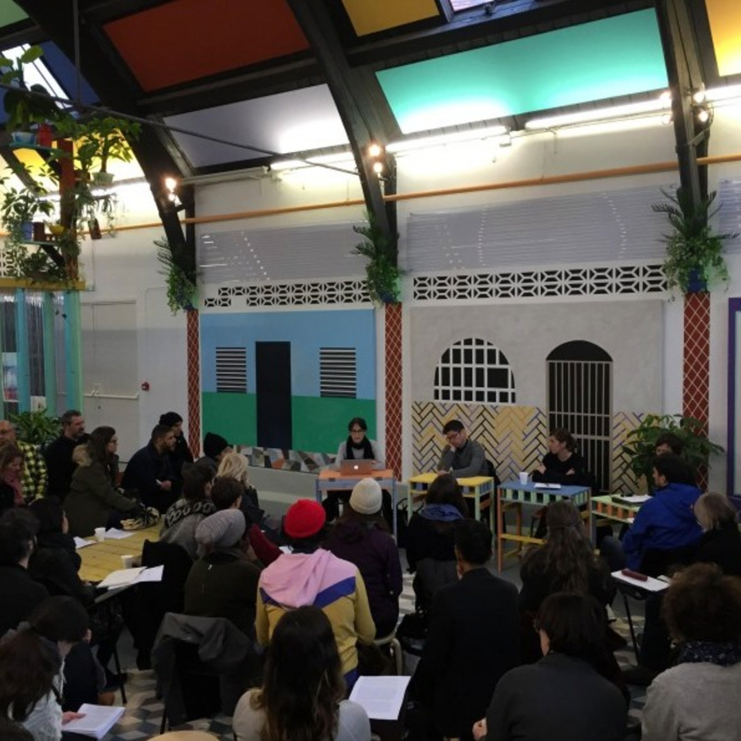 A group of people sit in a circle on stools inside a colourful, decorated room, attentively listening to a panel of speakers at the front. Some participants hold papers, and the atmosphere appears collaborative and engaged.